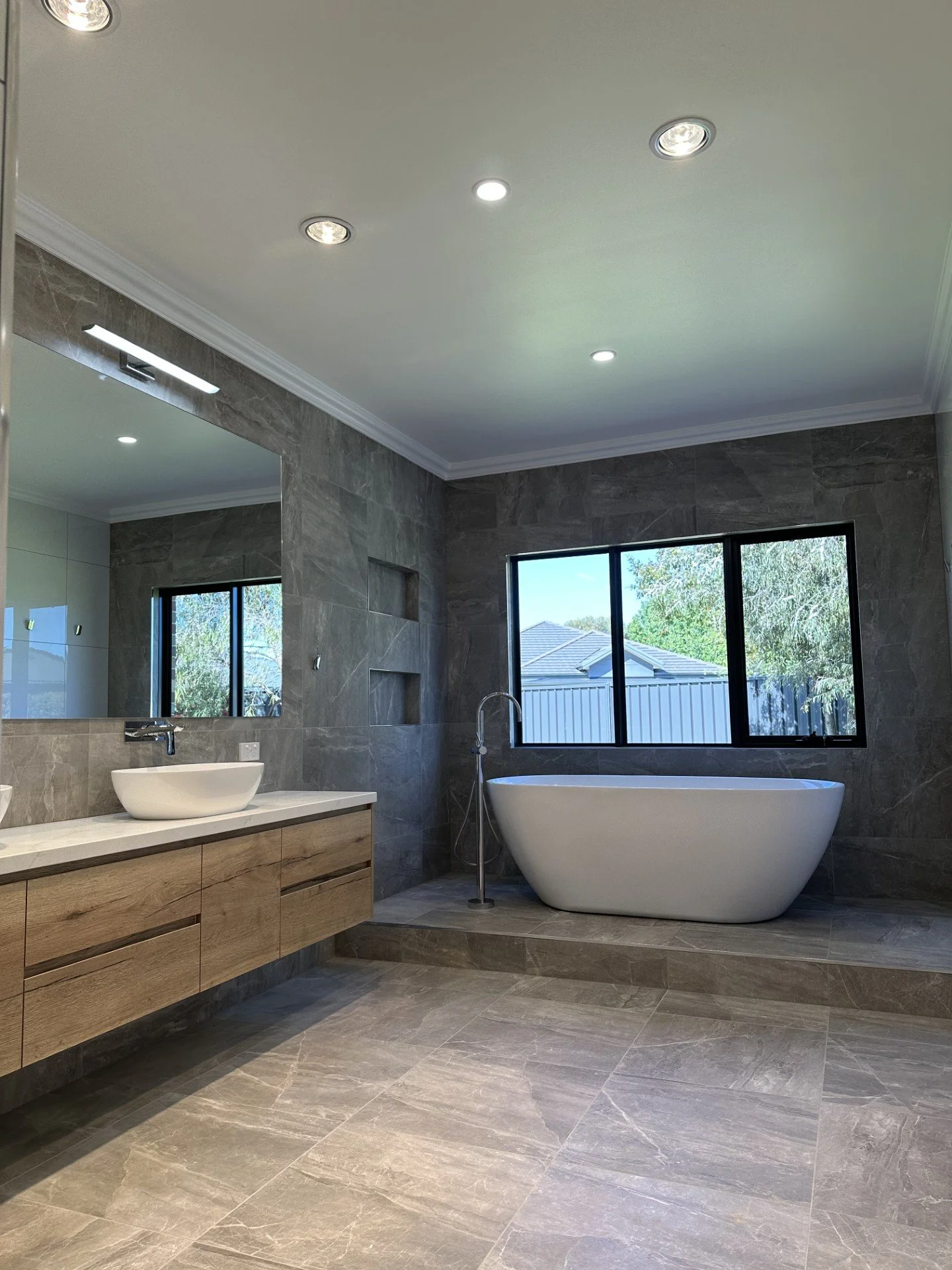 Modern bathroom featuring a freestanding bathtub, a large window showing outdoor greenery, a floating wooden vanity with a vessel sink, and a large mirror, all with gray stone tiles.