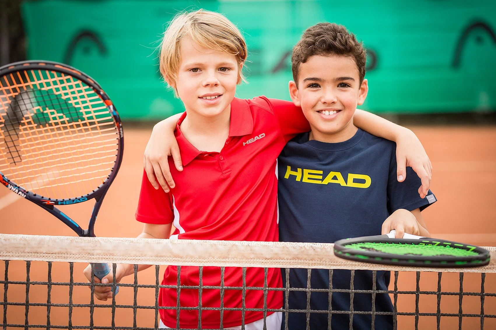 Two boys standing together on a tennis court, smiling with arms around each other's shoulders, holding tennis rackets.