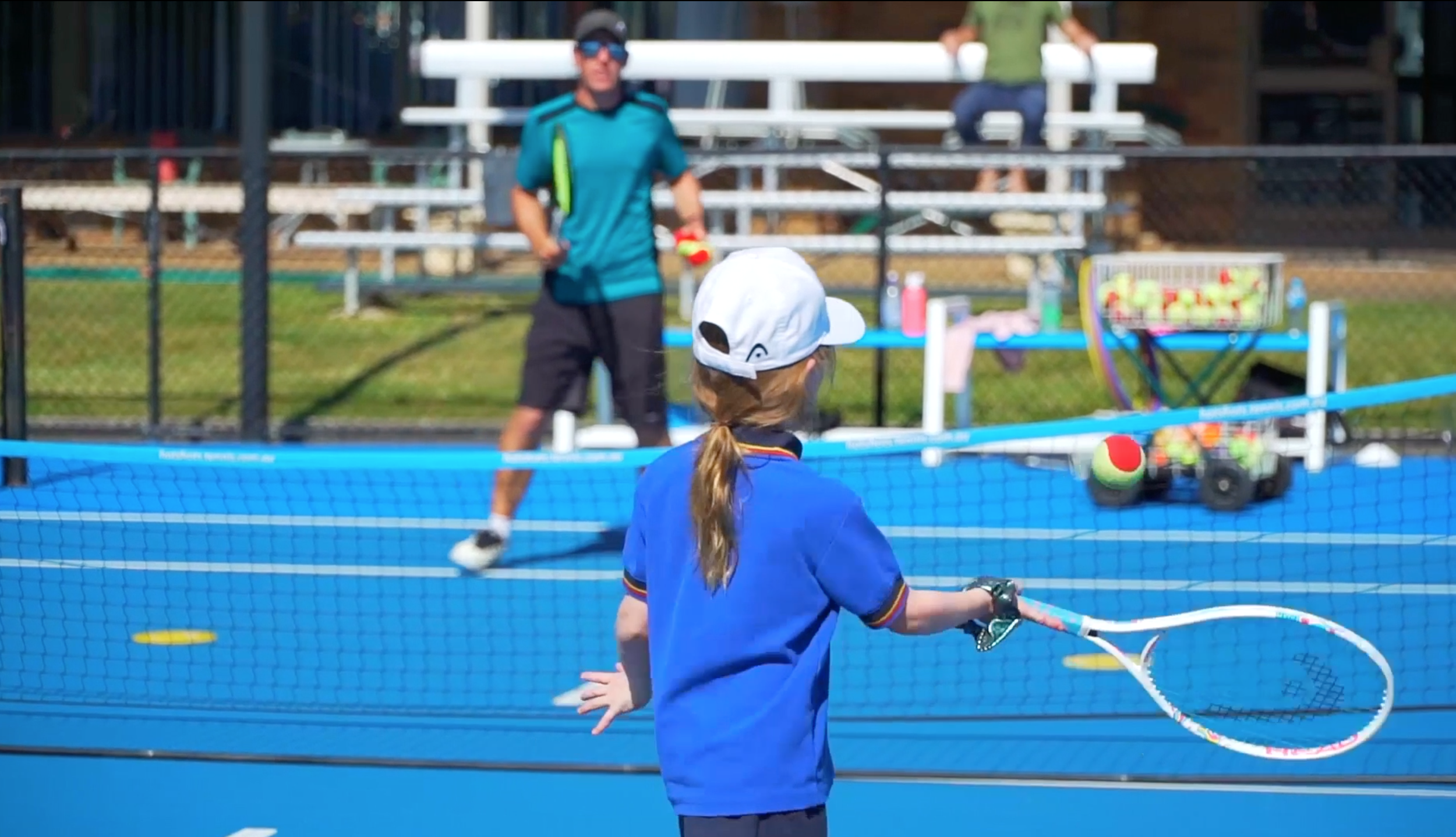 A young girl in a blue shirt and white cap playing tennis outdoors, with a coach in the background.