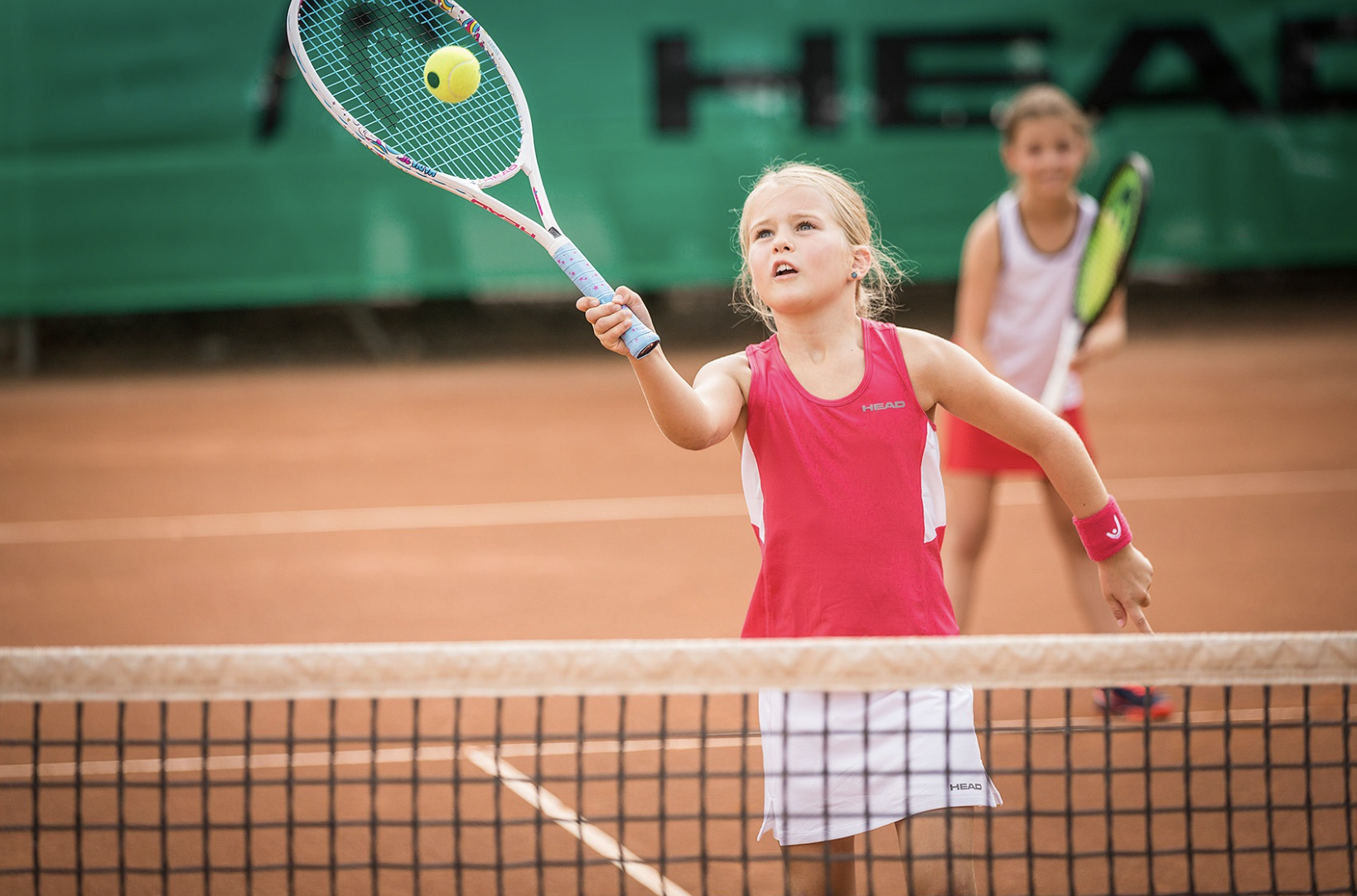 Two young girls playing tennis on clay court, one preparing to hit the ball with her racket.