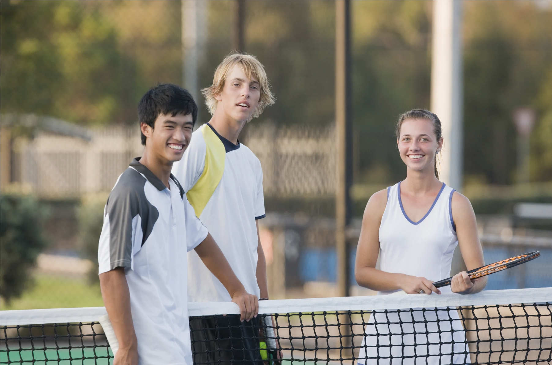 Three teenagers standing on a tennis court near the net, smiling, holding tennis rackets, with trees and a fence in the background.