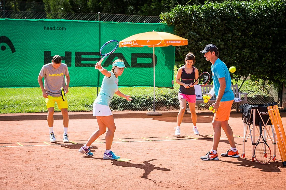 A tennis lesson on a clay court with a coach and three students. The coach is teaching how to serve, with tennis balls and rackets.