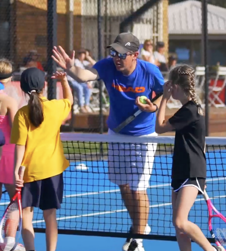 Tennis coach giving high five to young girl at net during practice, with other children in background