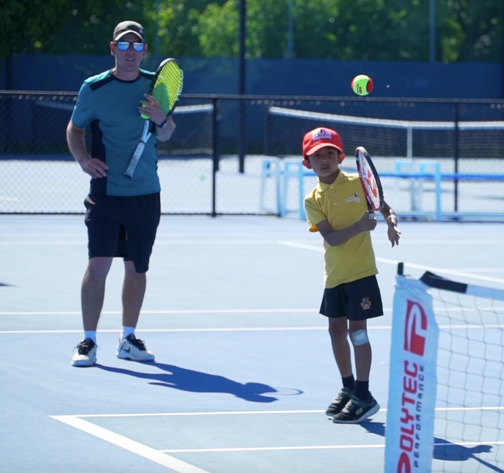 A man and a young boy playing tennis on an outdoor court. The man is wearing sunglasses, a blue shirt, black shorts, and white sneakers. The boy is wearing a red cap, yellow shirt, black shorts, and tennis shoes. The boy is preparing to hit a tennis ball with a racket.