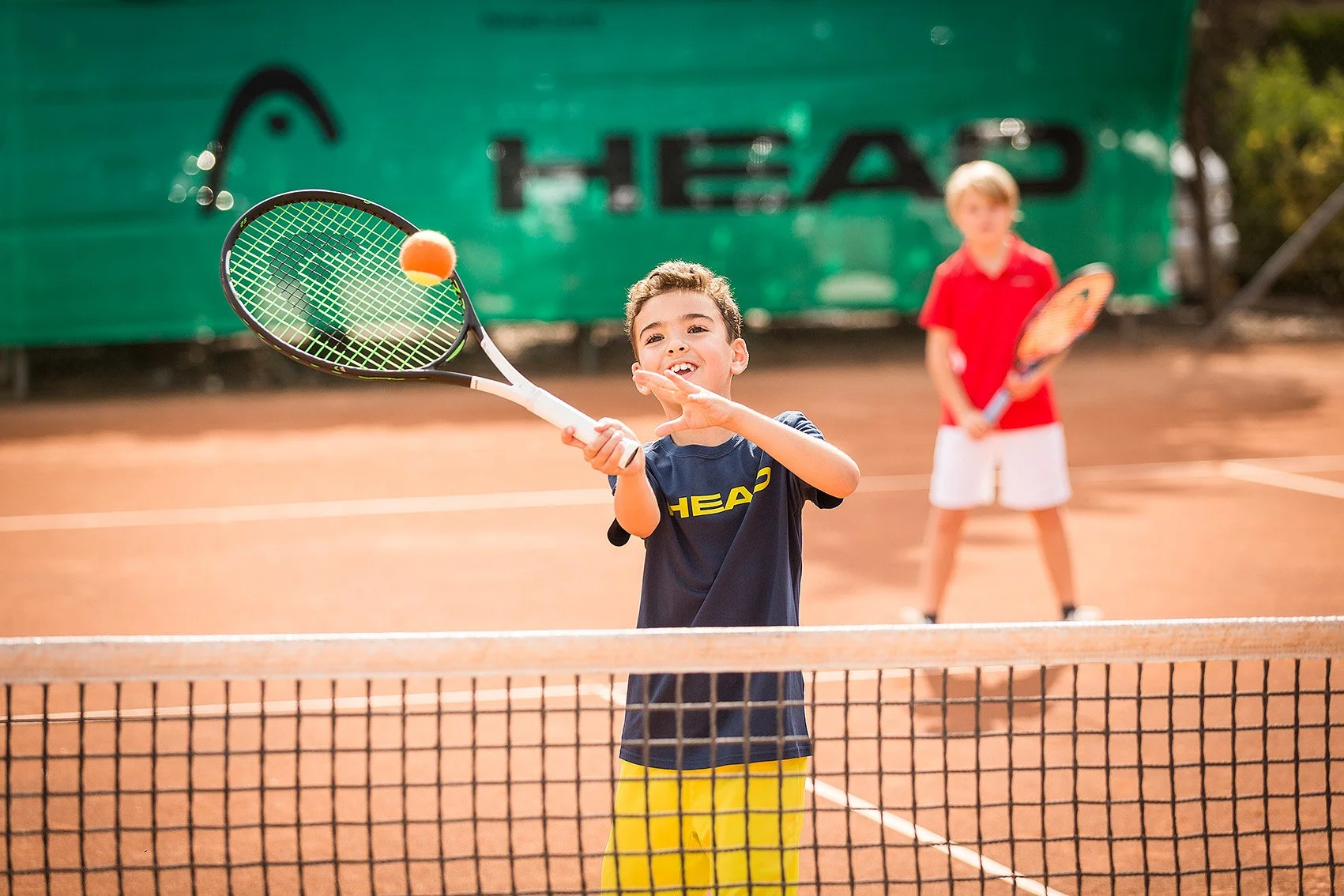 Two young boys playing tennis on an outdoor clay court. One boy is hitting a tennis ball with a racket, smiling, in the foreground. The other boy, in the background, is holding a tennis racket and watching. There is a green hedge with a large black and green banner with the word "HEAD" behind them.