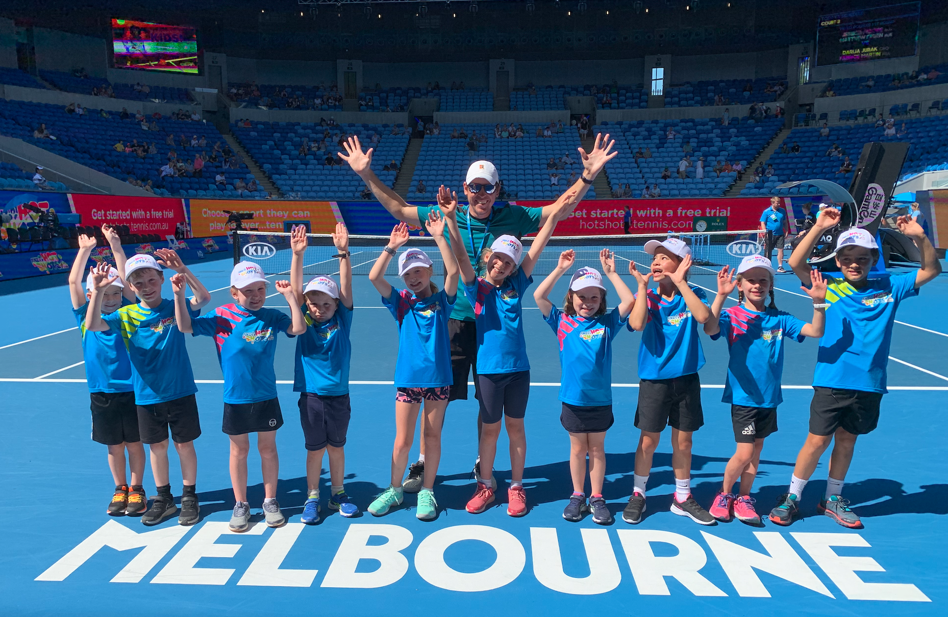 Group of children and their coach on a tennis court at the Melbourne Park during Australian Open, wearing blue shirts and white caps, smiling and raising their hands.