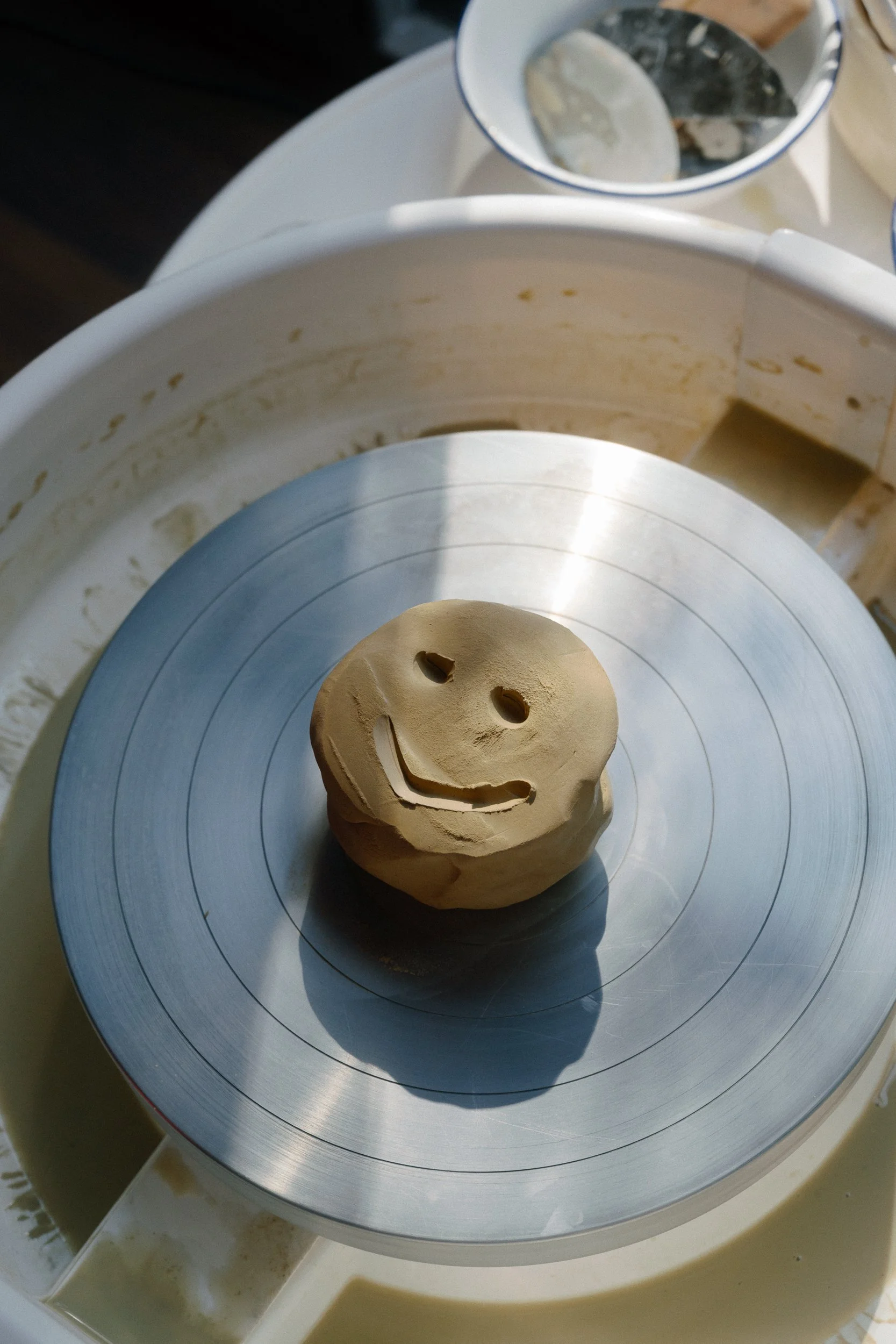 A smiley face is carved into a ball of yellow clay on a pottery wheel.
