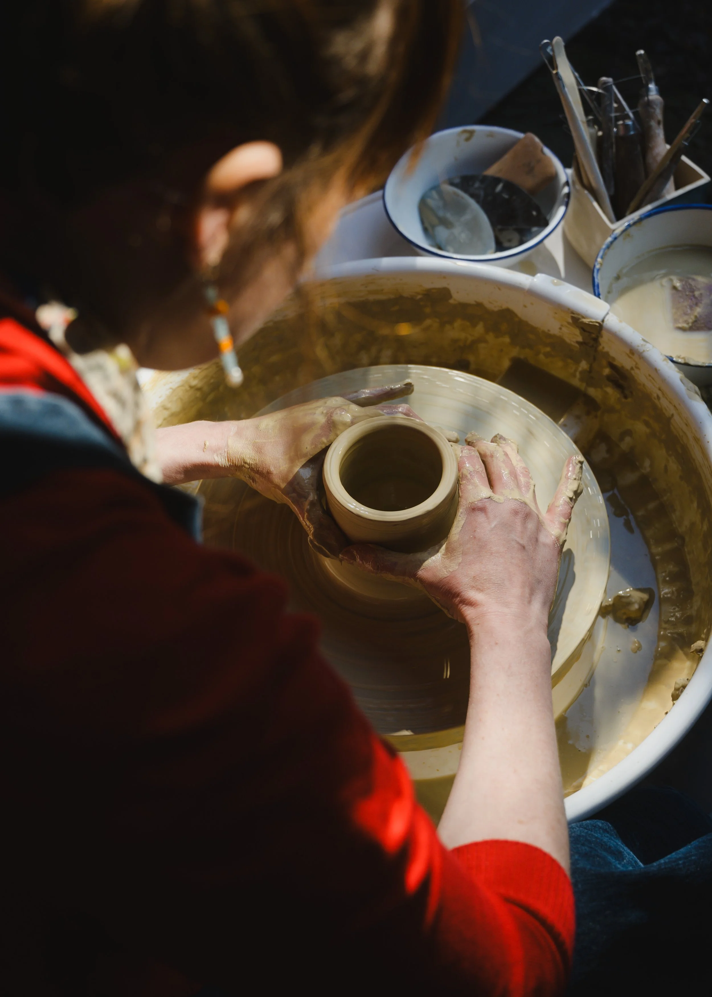 Person shaping a clay pot on a pottery wheel with various pottery tools nearby.