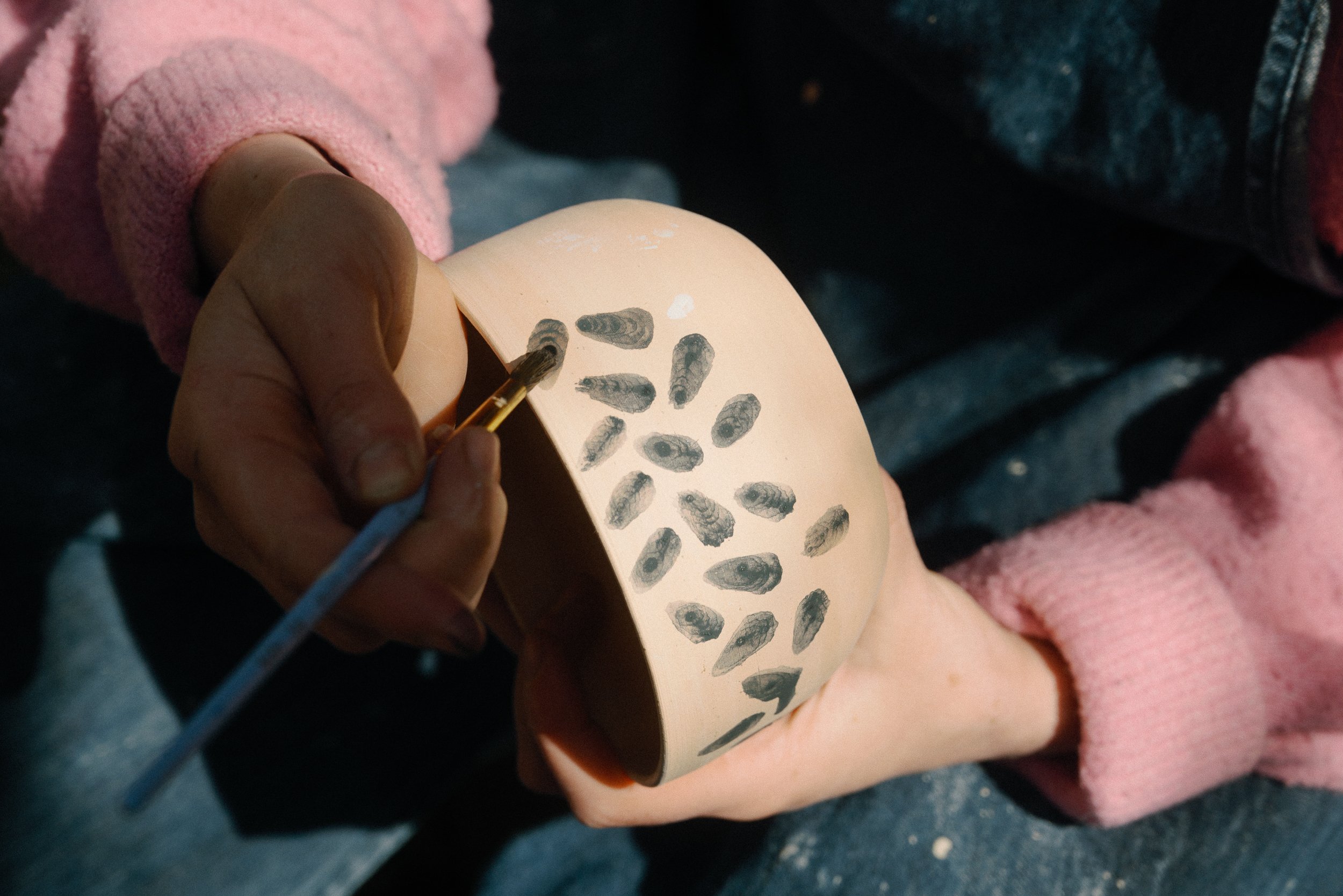 A person in a pink sweater is decorating a ceramic bowl with black ink in the shape of clam shells using a small paintbrush.