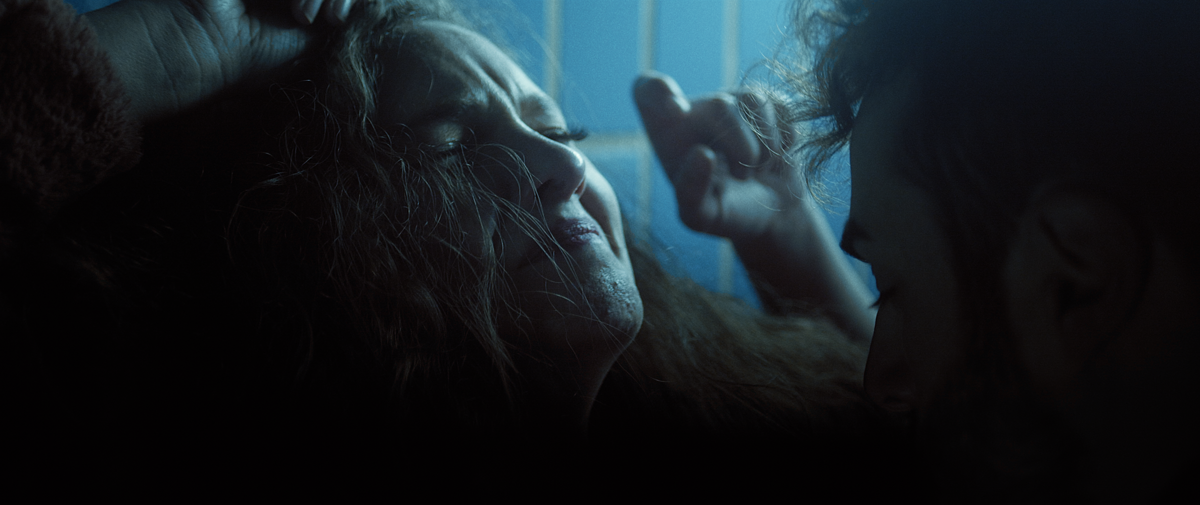 A woman with long, curly hair lying down with her eyes closed, and a man with curly hair leaning close to her, touching her face gently in a dimly lit scene.