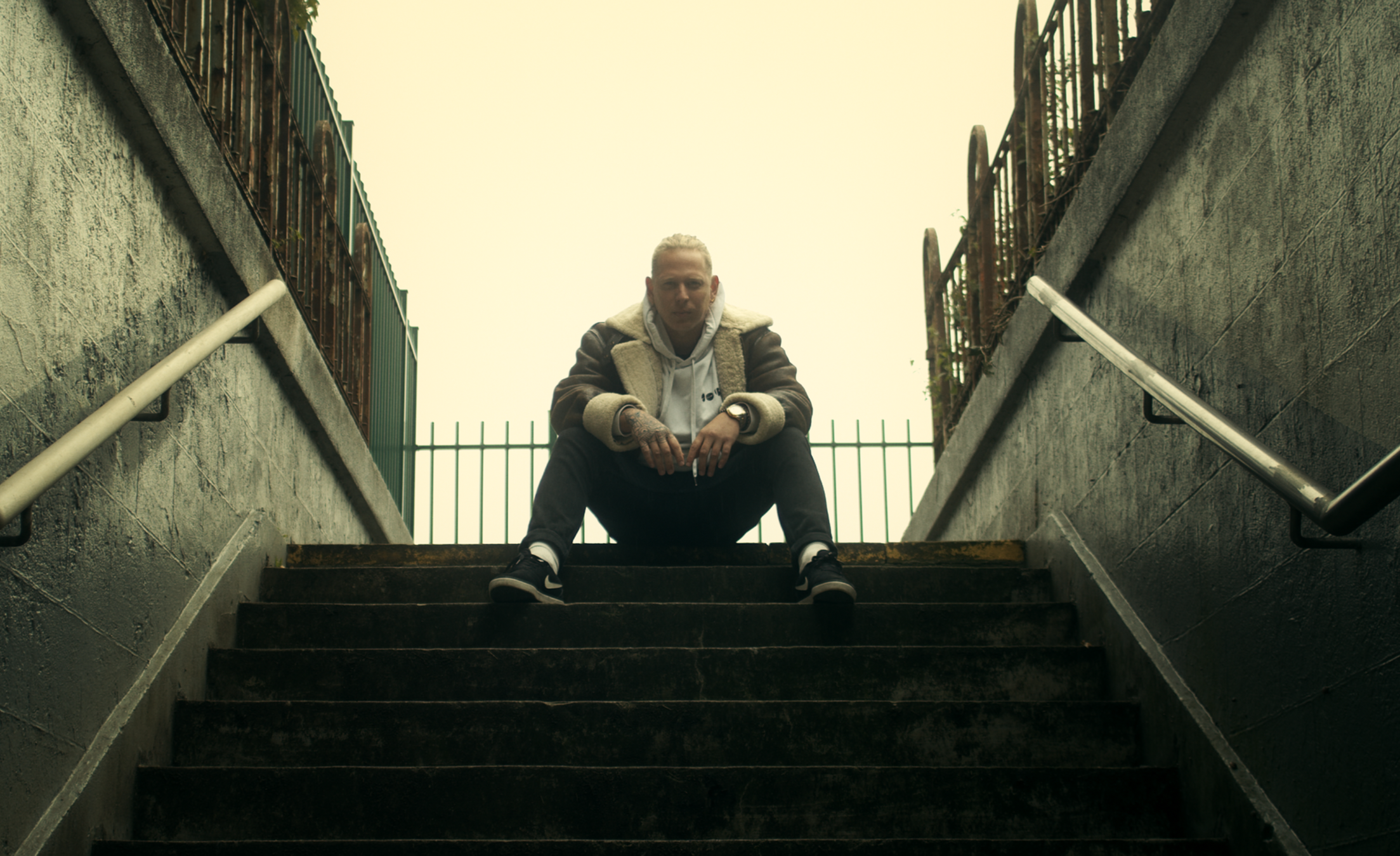 A young man sitting on stairs in an urban setting, with fences and railings on either side, looking directly at the camera.