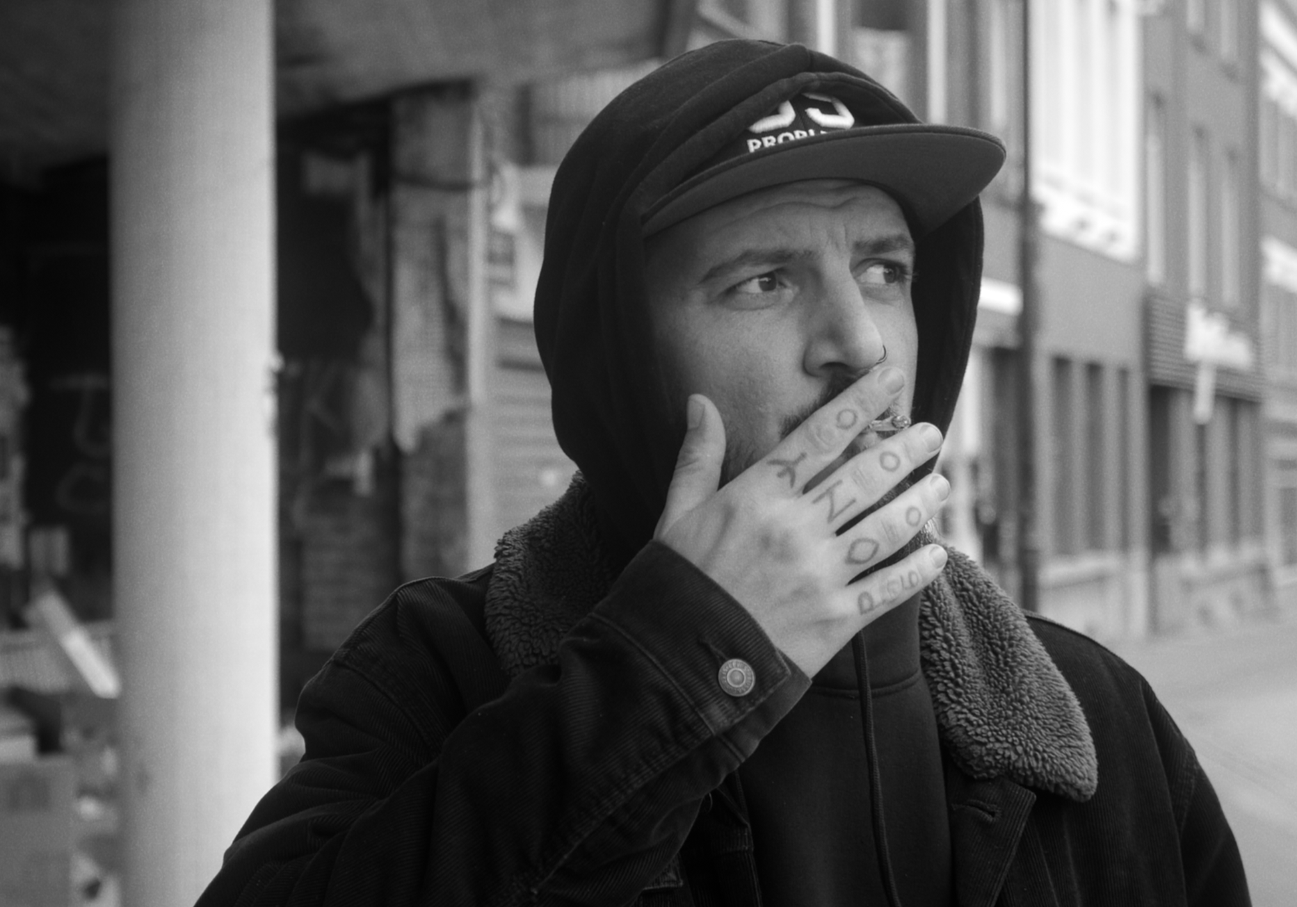 A young man with a beard and tattoos on his fingers, wearing a cap, hoodie, and jacket, stands outdoors and appears to be smoking in an urban environment.