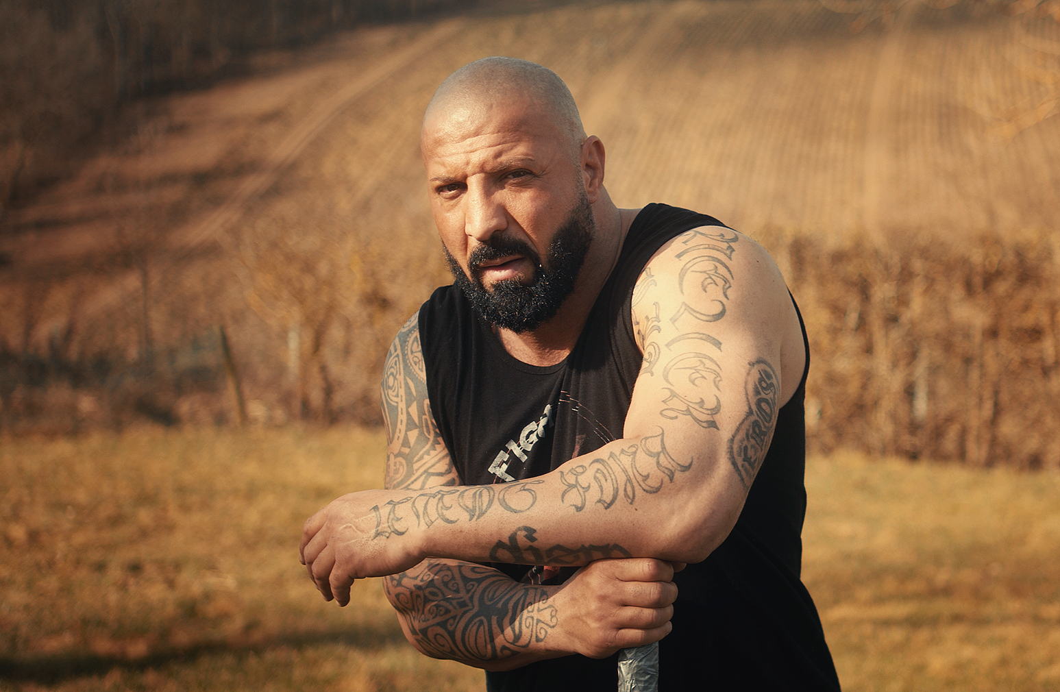A muscular man with a shaved head and beard, covered in tattoos, standing outdoors in a field of dry grass on a sunny day.