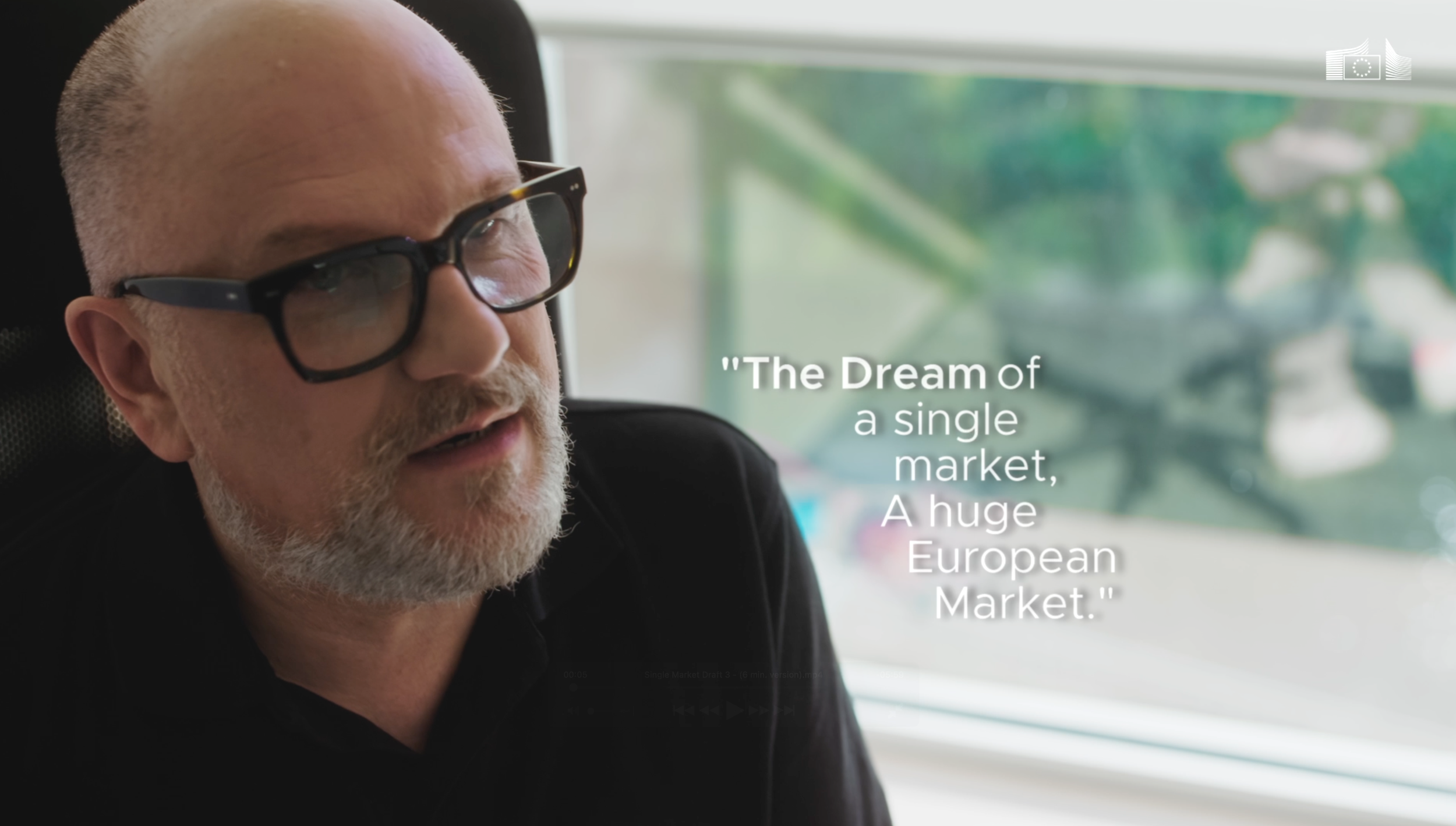 Man with gray beard, glasses, and short gray hair sitting in a car with a window view of trees, with quote about European market on the side.