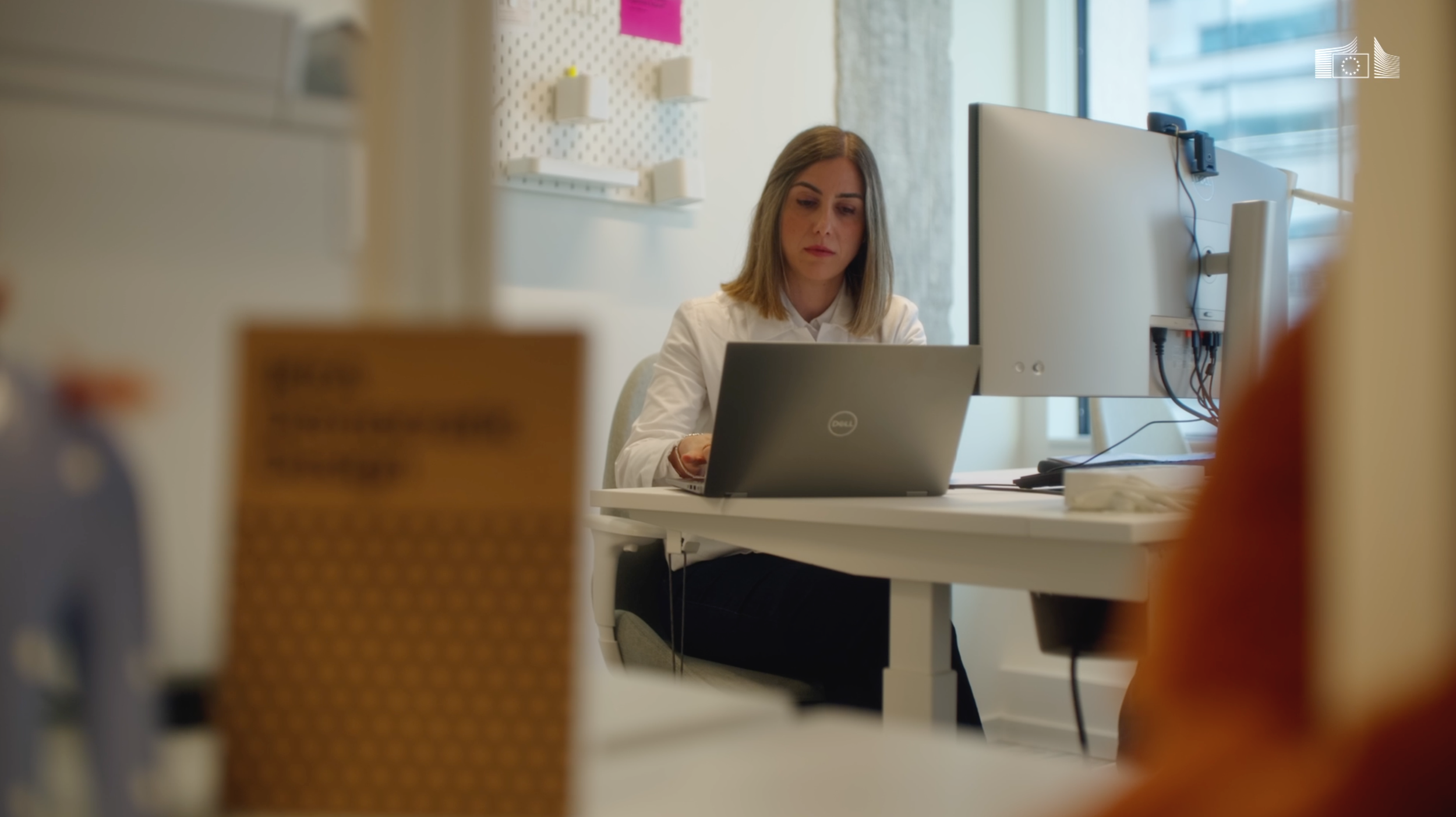 A woman working on a laptop at a desk in an office, with a large monitor and window in the background.