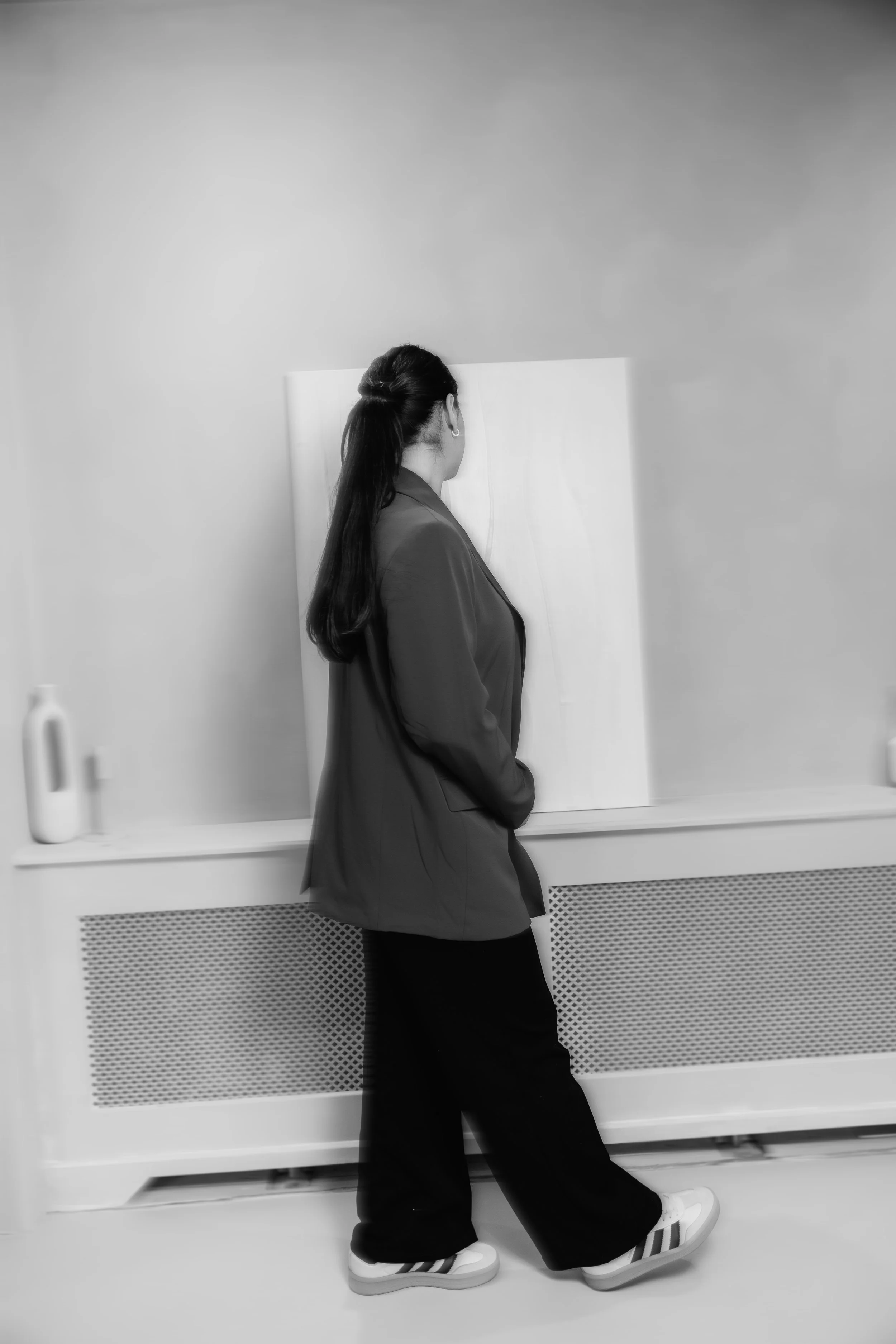 Een vrouw met lang haar draagt een blazer en brede broek, staat naast een radiator en kijkt naar een leeg wit canvas op een muur.