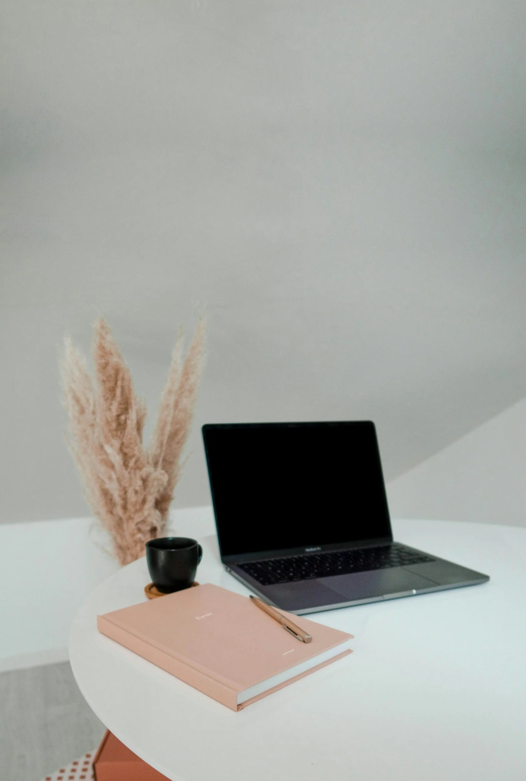 Werkplek met een laptop, een roze notitieboek, een pen, een zwarte kop koffie, en een vaas met gedroogd pampasgras.