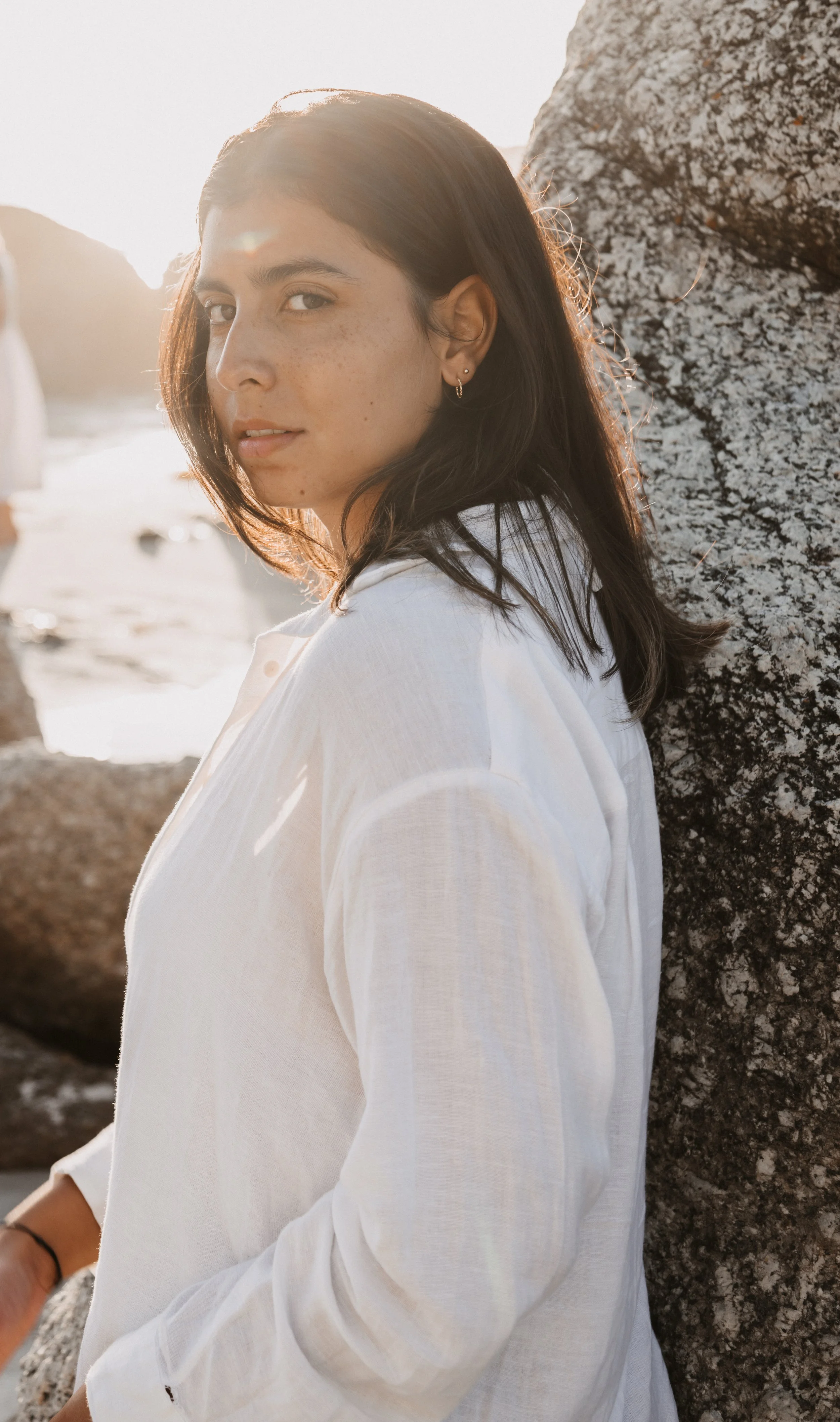 Vrouw met donker haar en lichte huid, zittend buiten bij zonsondergang, met grote rotsen op de achtergrond.