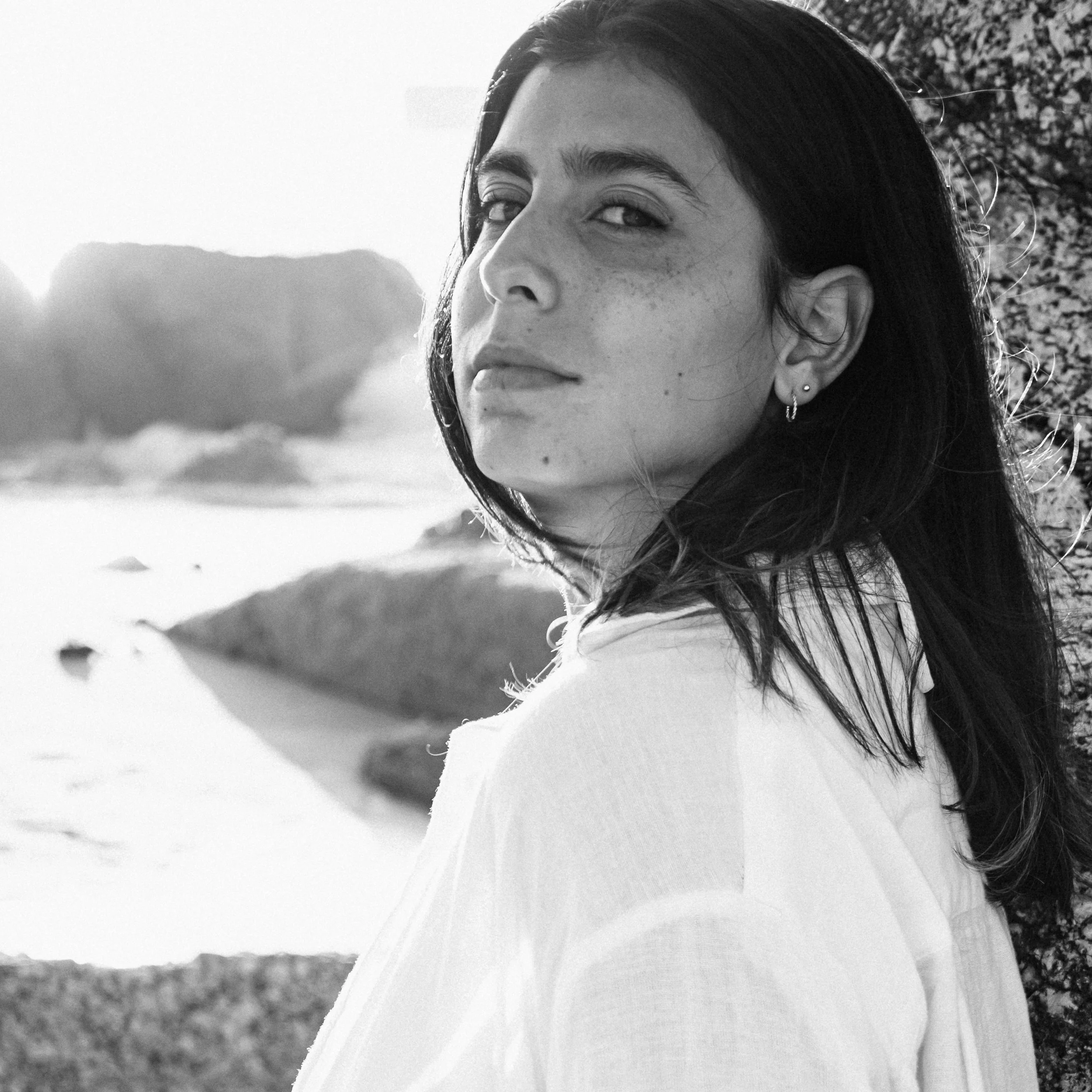 Portret van een vrouw met zwart haar, lichte huid en sproeten, kijkend naar de camera, met een natuurlijke achtergrond.