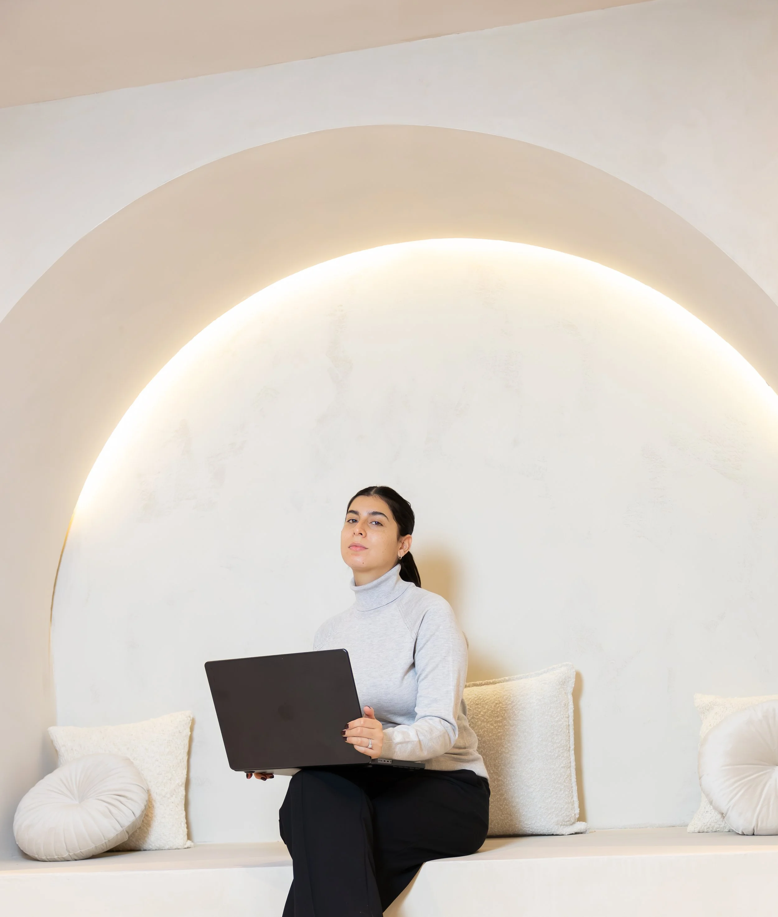 Vrouw met een laptop zittend op een wit bankje met kussens, in een modern, minimalistisch interieur.