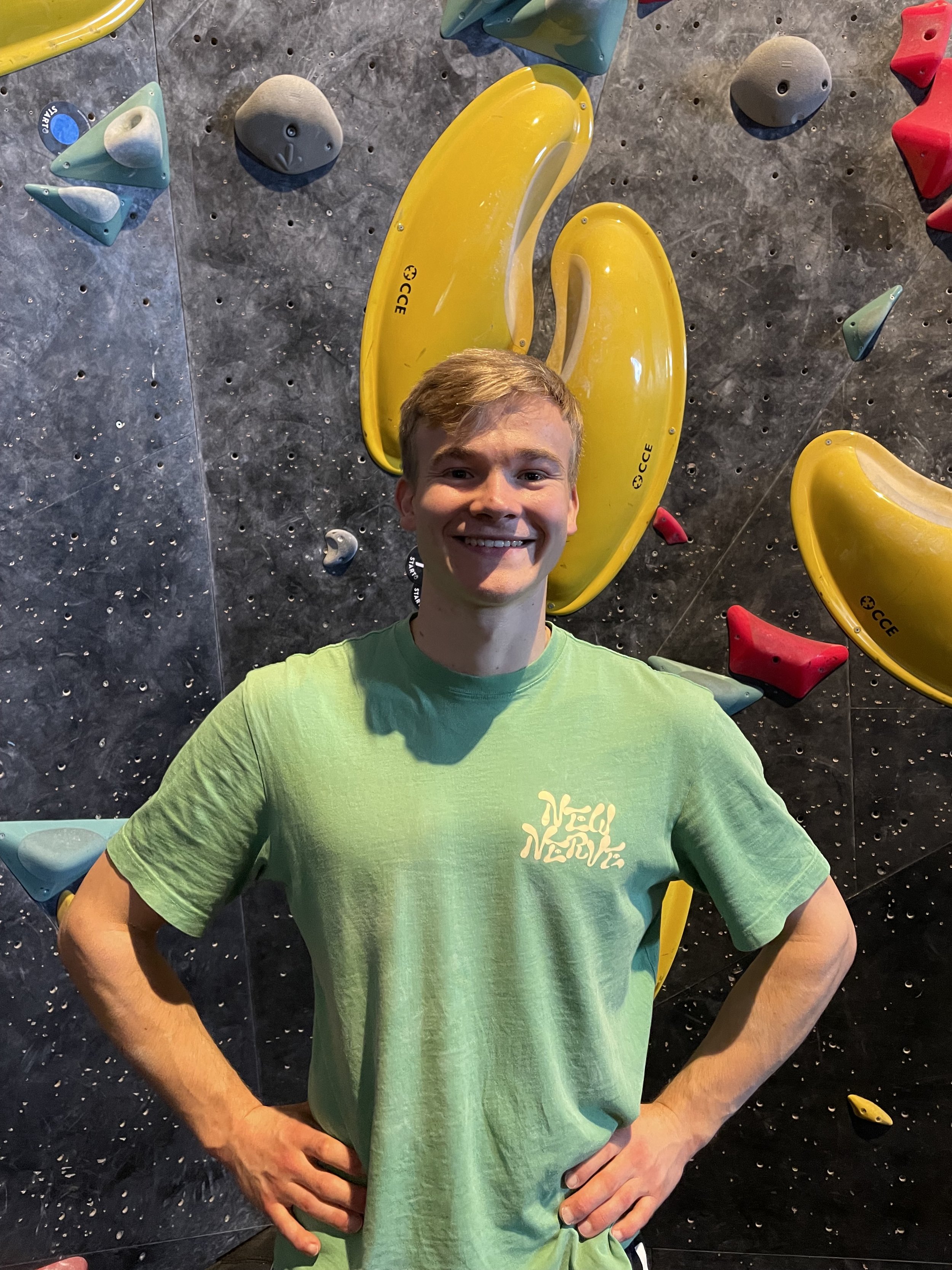 Junger Mann mit kurzen blondem Haar, grünem T-Shirt, lächelnd, steht vor einer Kletterwand mit bunten Griffen.