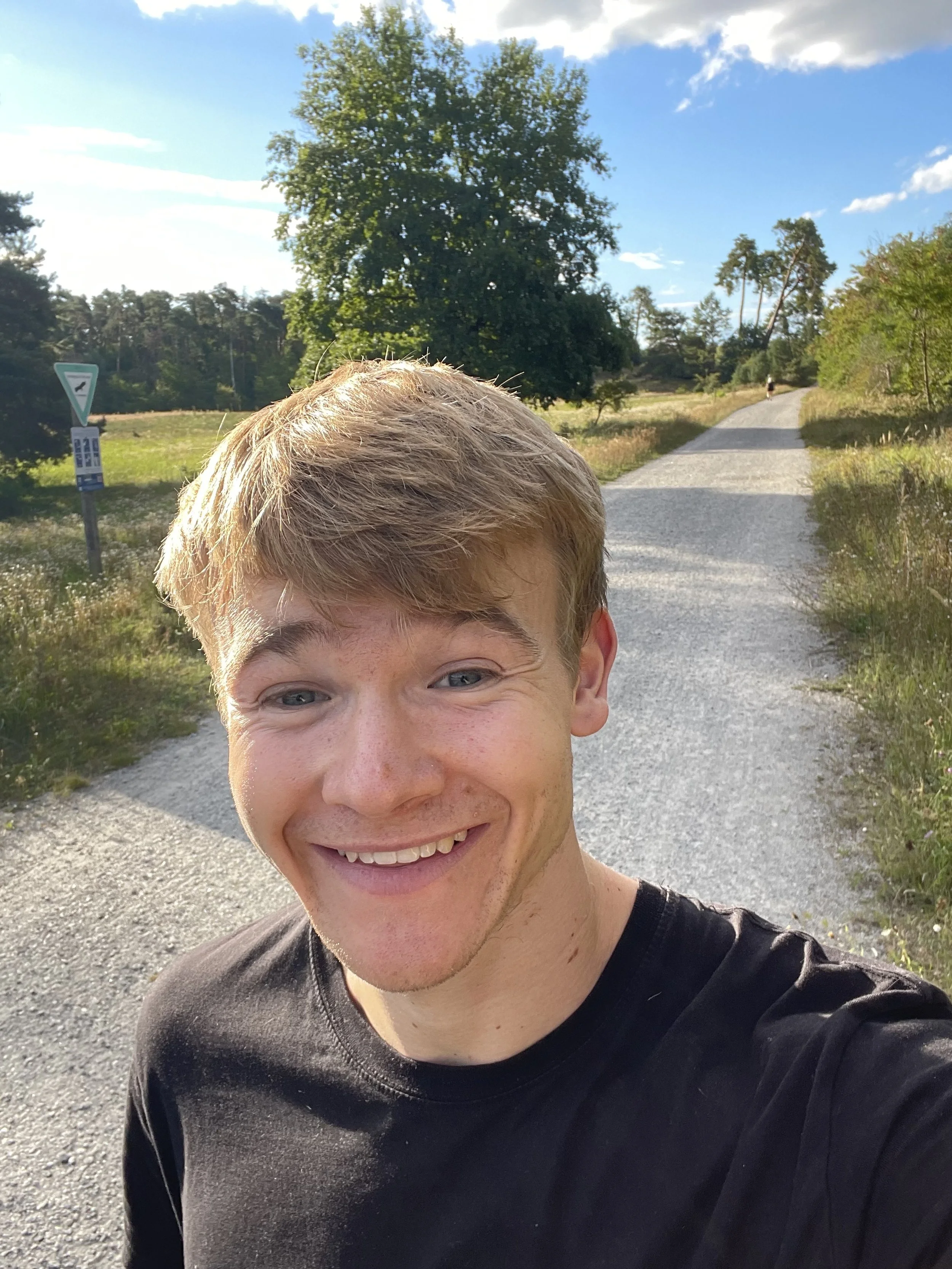 Ein junger Mann macht einen Selfie auf einem Feldweg bei sonnigem Wetter, umgeben von Bäumen und hohen Gräsern.