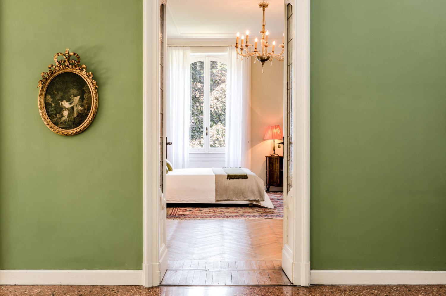 Vista di una stanza da letto attraverso una porta aperta. La stanza ha pareti color pesca, un grande finestrone con tende bianche, un letto con copertura beige, una lampada rosa, un comodino in legno e un lampadario chandelier.
