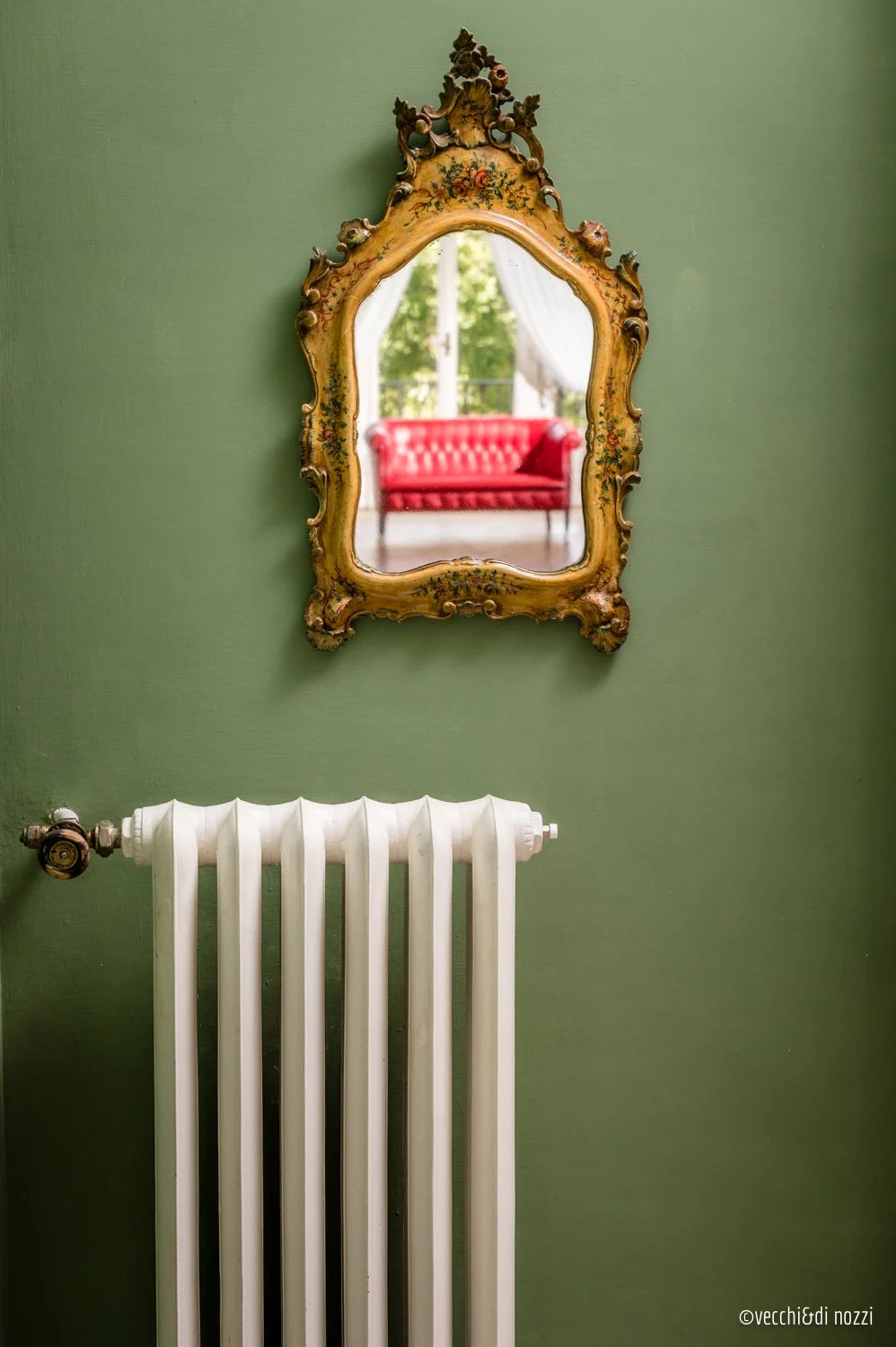 Specchio antico con cornice decorata appeso su parete verde, sotto termosifone bianco, riflesso di una finestra con tende e divano rosso.