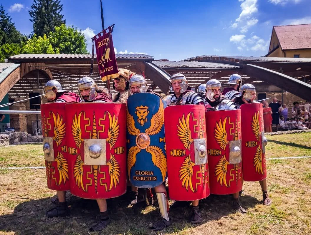 A group of people dressed as Roman soldiers, wearing helmets and armor, holding large red shields with gold designs, standing outdoors with a small building and trees in the background.