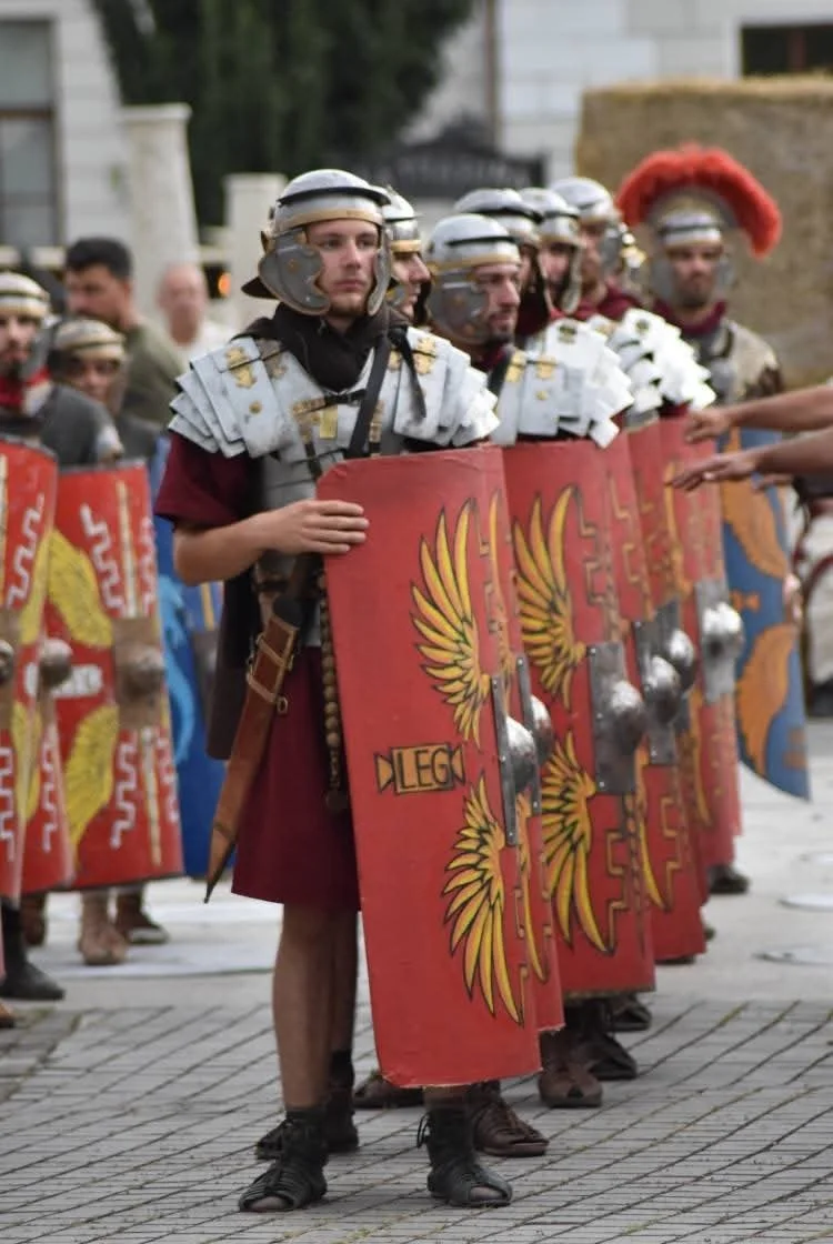 Group of people dressed as Roman soldiers standing in a row, holding large rectangular shields painted with yellow and red designs, outdoors in front of stone buildings.