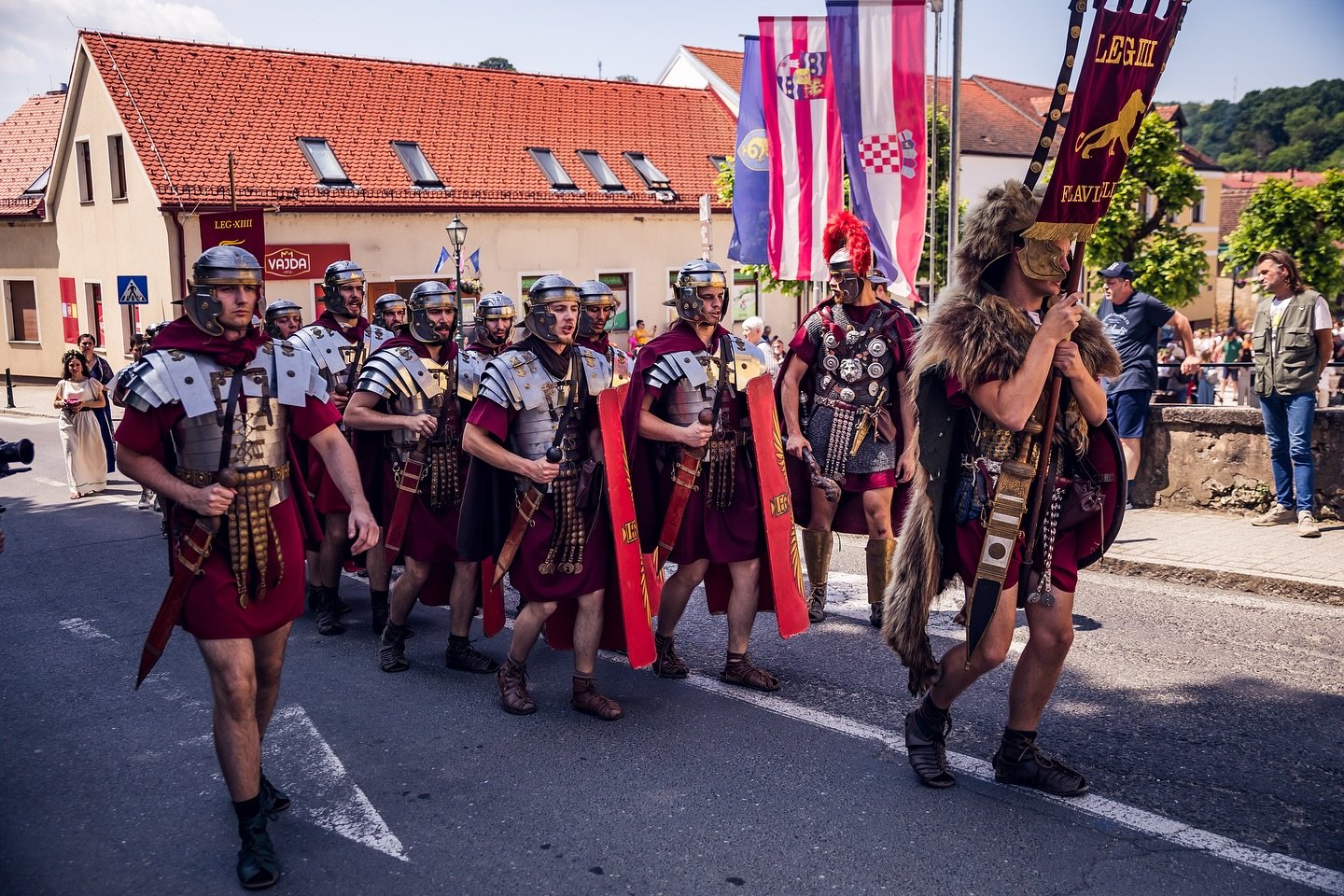 Group of people dressed in Roman and medieval warrior costumes participating in a parade or reenactment on a street in a small town, carrying shields, banners, and flags.