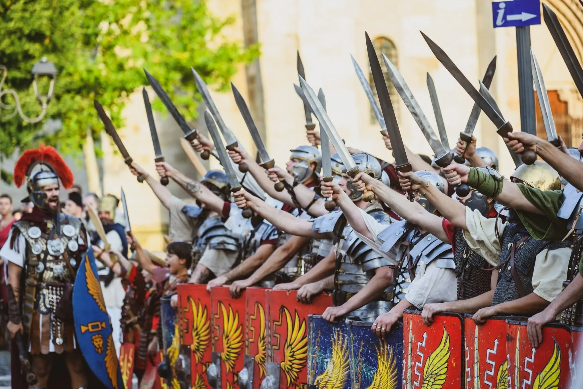 A reenactment of a Roman battle, with participants dressed in Roman armor, helmets, and shields, holding swords, and standing in formation against a backdrop of trees and historical buildings.