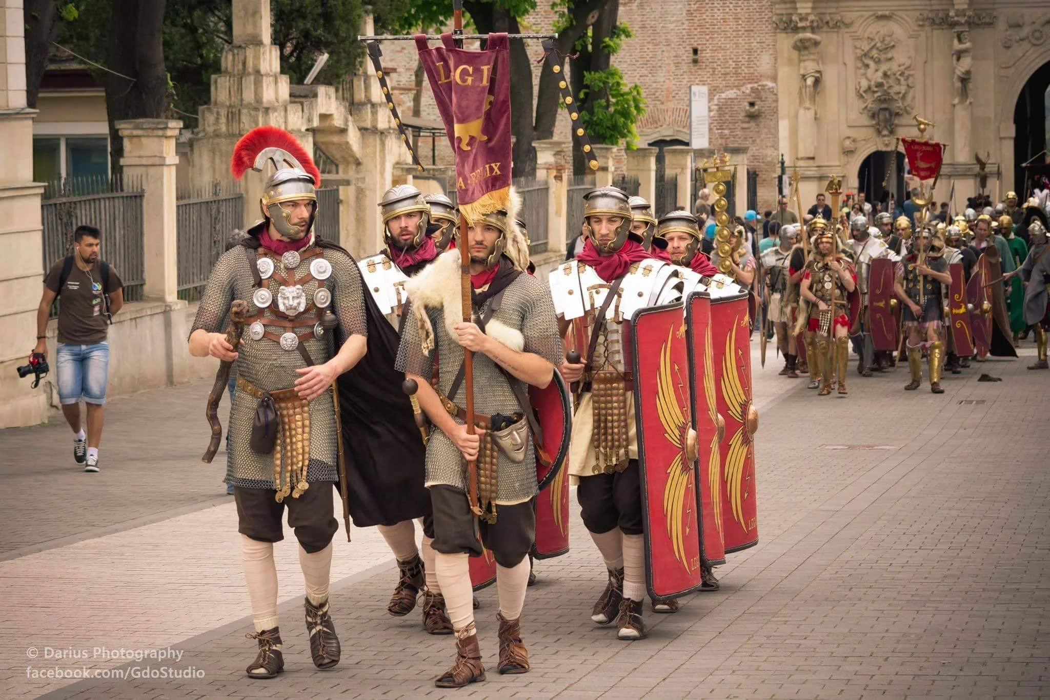 A group of men dressed as Roman soldiers in a historical reenactment, walking in a parade on a city street with spectators watching.