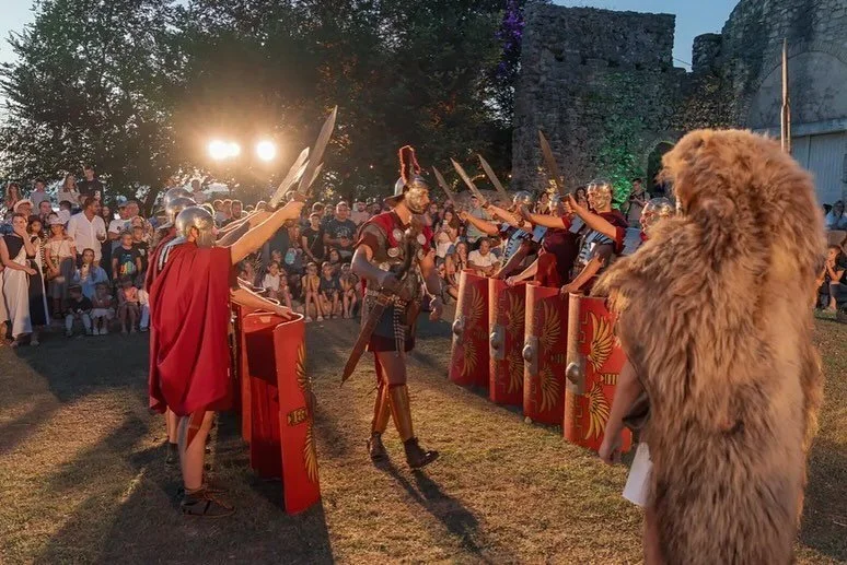 A reenactment of a Roman gladiator confrontation with spectators watching, some dressed in ancient Roman costumes and a large person in a bear costume, outdoors during sunset.