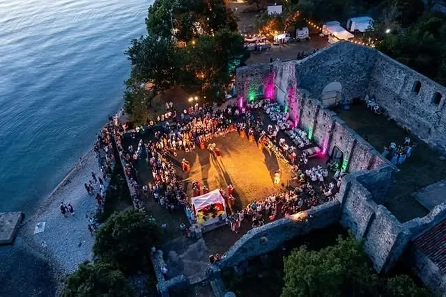 A nighttime outdoor wedding ceremony inside the ruins of an old stone building near a body of water. Guests are seated around a central aisle, with colorful lights illuminating part of the structure.