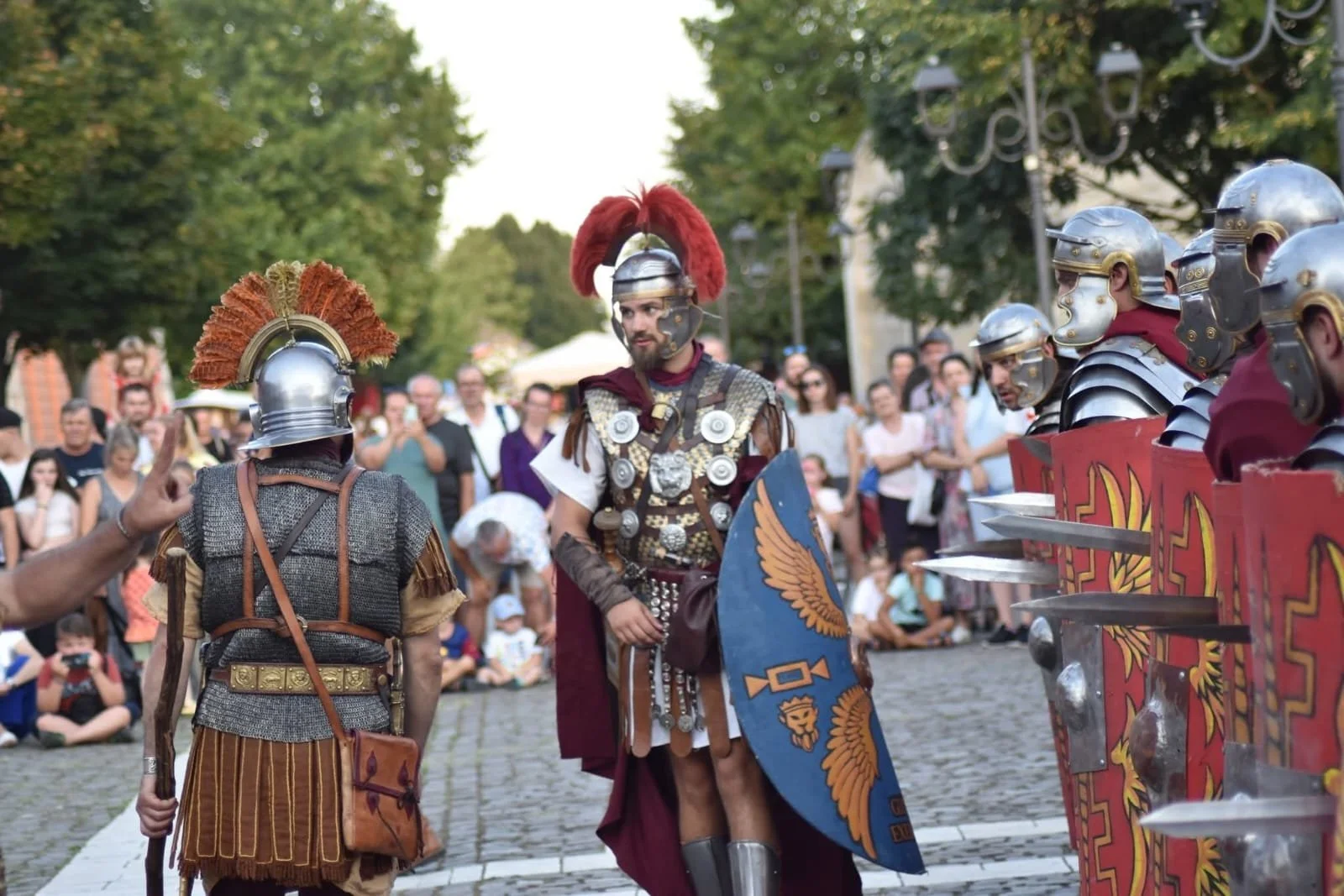 Roman soldiers in armor and helmets with plumes, carrying shields and swords, standing in front of an audience during a historical reenactment.