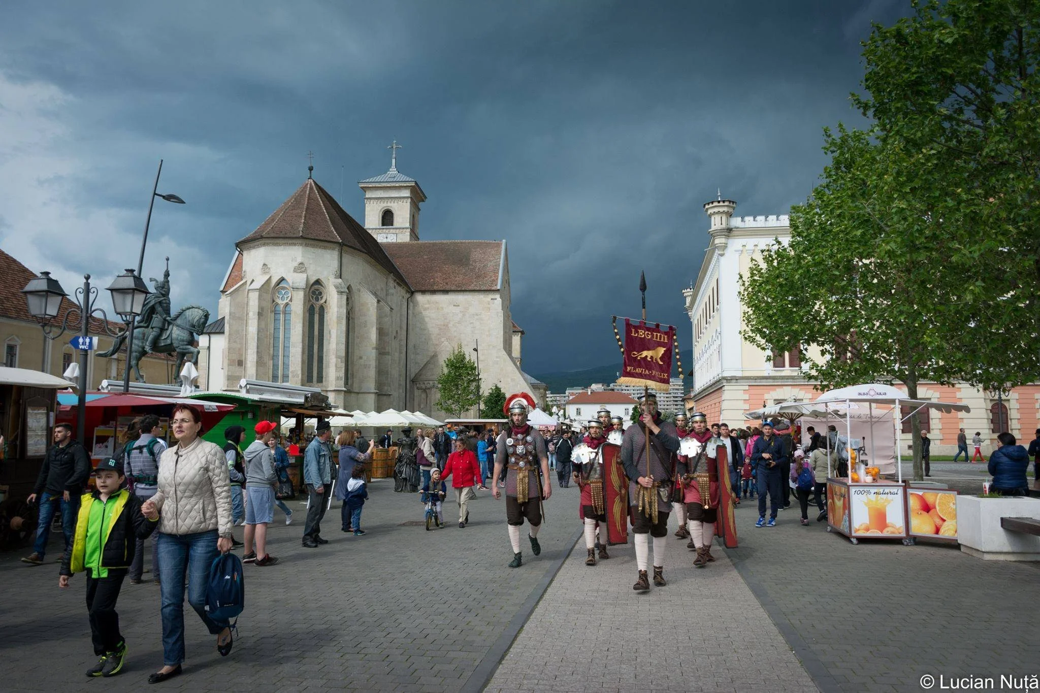 A crowd in a town square under dark stormy clouds, with a marching group of people dressed as Roman soldiers carrying a banner, surrounded by market stalls, a statue of a mounted knight, and historic buildings.