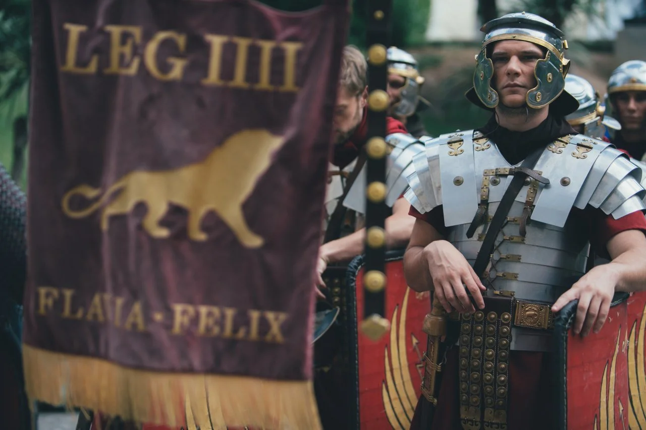 A man dressed as a Roman soldier standing with others in the background, holding a shield, during a historical reenactment event.