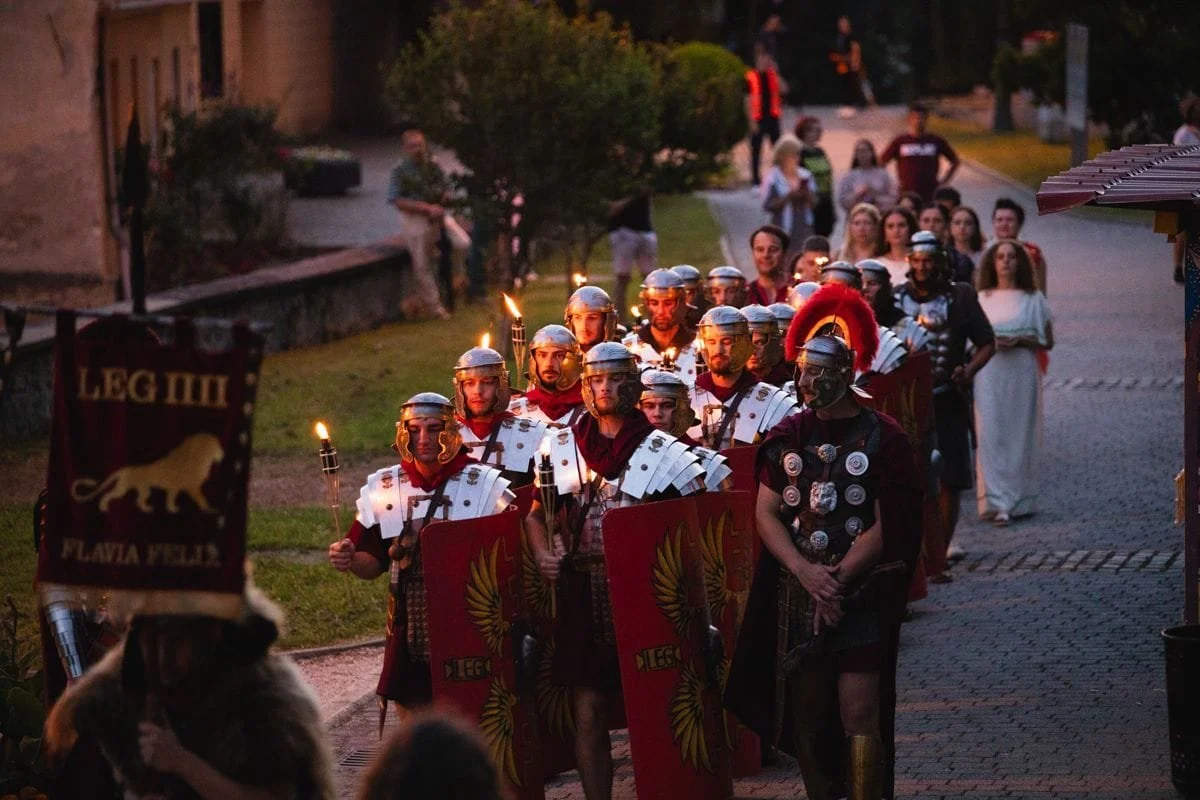 A Roman-themed parade or reenactment with people dressed as Roman soldiers, carrying shields and torches, walking down a street at dusk, with onlookers watching along the sidewalk.