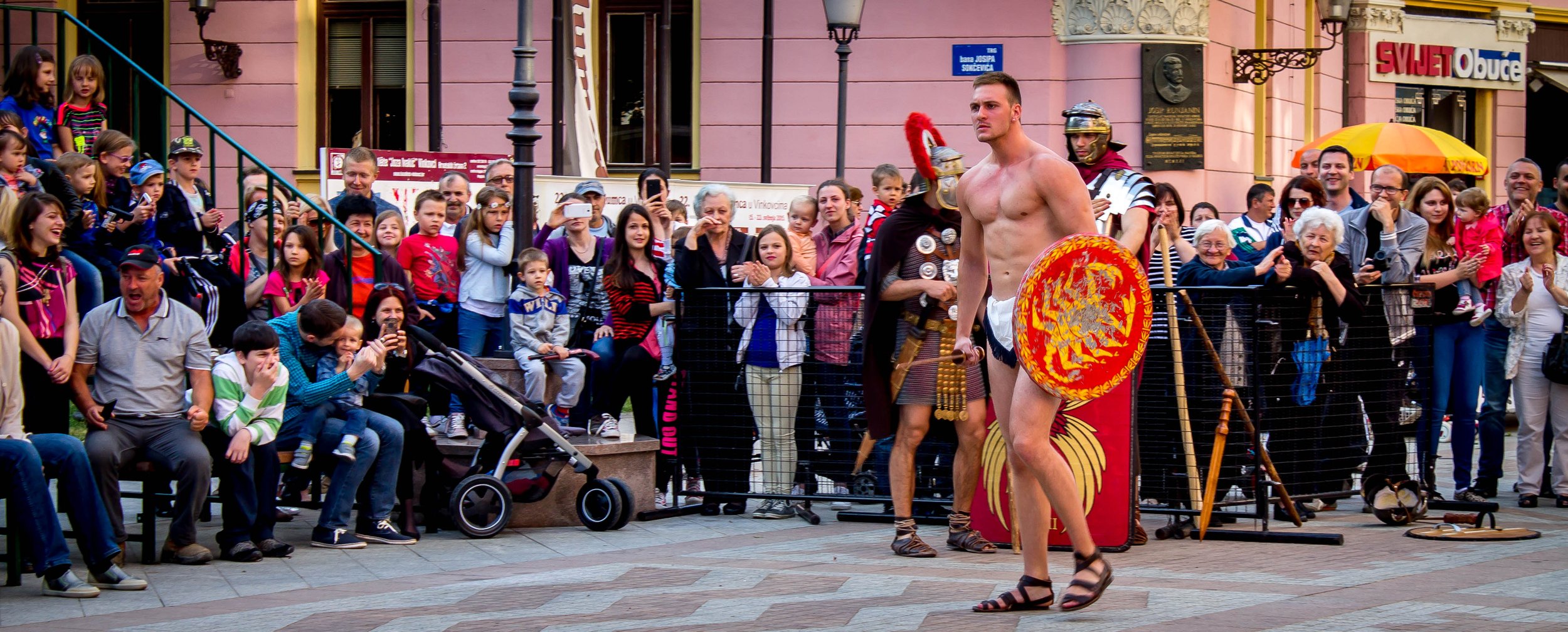A street performer dressed as an ancient Roman soldier, holding a shield, stands in front of a crowd of children and adults watching a historical reenactment or show in an urban setting.