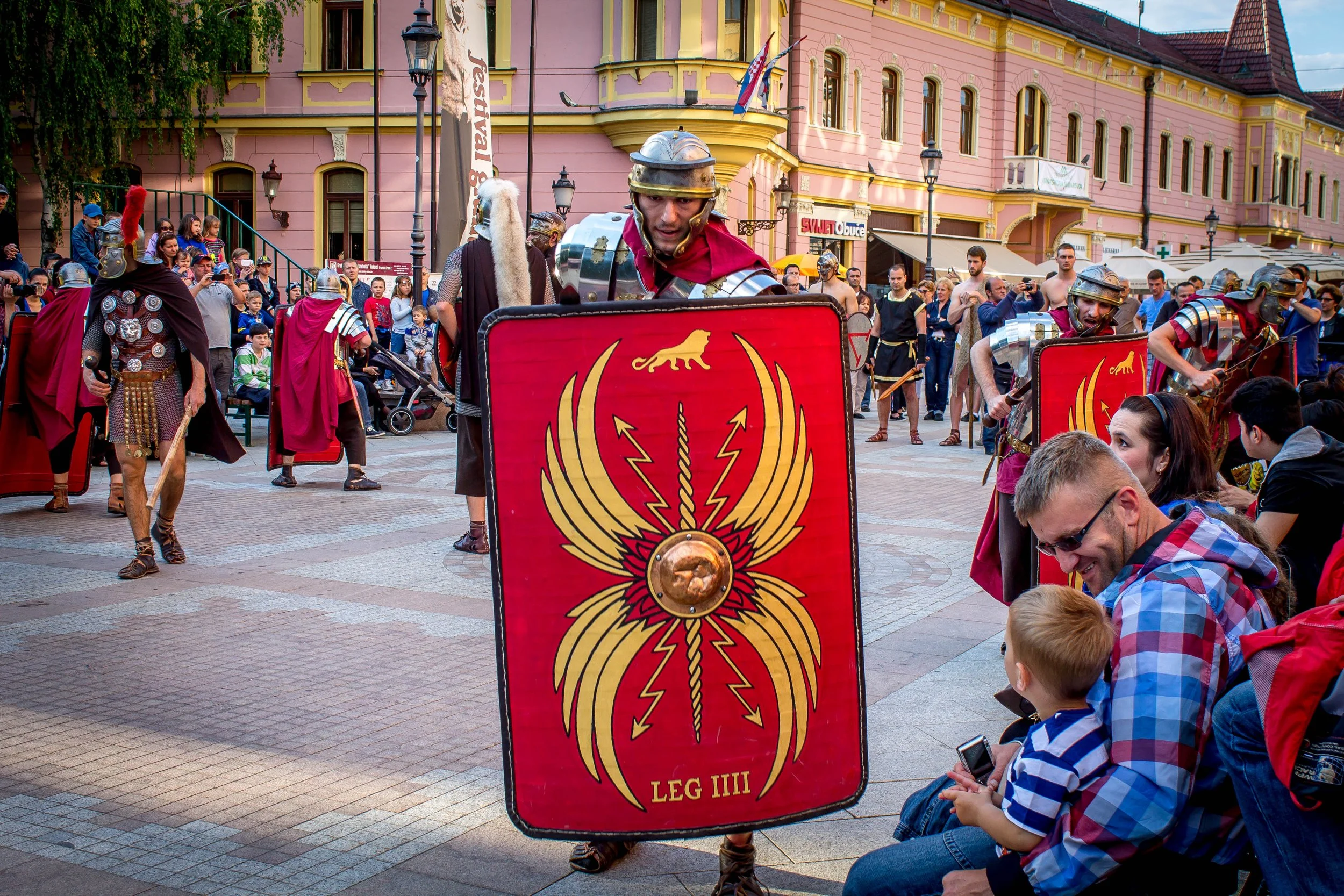 Reenactors dressed as Roman soldiers with shields in a public square during a festival, with onlookers sitting and watching.