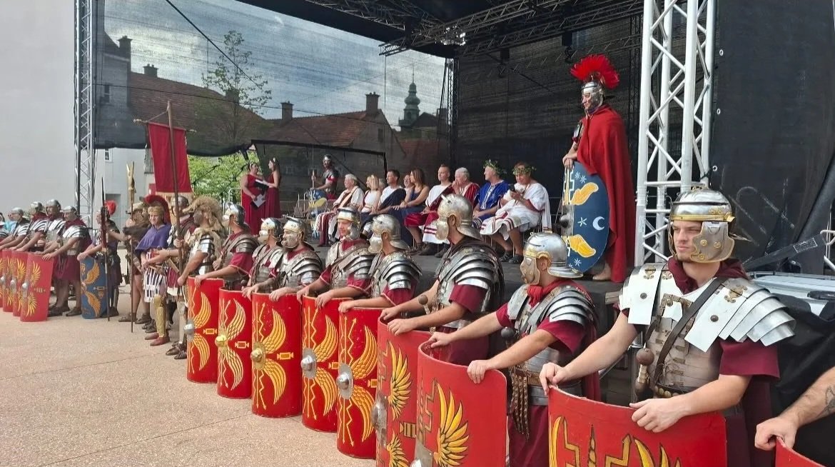 A group of people dressed as Roman soldiers with shields and helmets are standing in a line, facing forward. Behind them, there is a stage with seated individuals, some dressed in historical or Roman-style costumes. The stage has a person dressed as a Roman senator or emperor, wearing a red cape and a crest with a helmet, standing in front of a shield decorated with a blue background, yellow fish, and crescent moons. The scene appears to be part of a historical reenactment or cultural event.