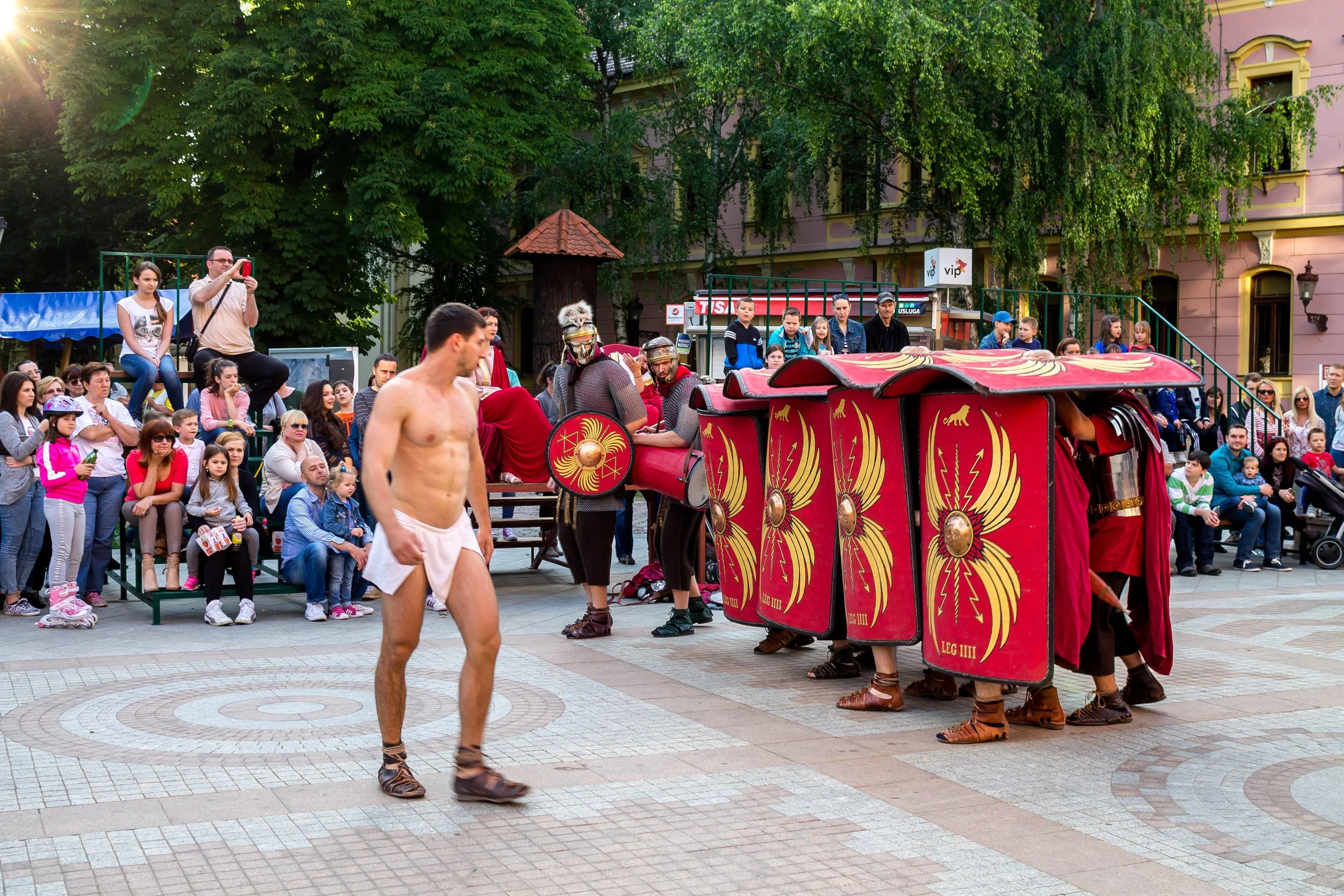 A street performer dressed as an ancient Roman soldier without a shirt and in a white towel, walking past a Roman-style military reenactment with soldiers in armor holding red shields and a red chariot, while a crowd of spectators watches.