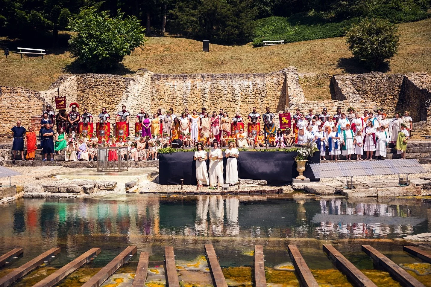 A large group of people dressed in ancient Roman attire, including tunics and armor, performing a reenactment or show by a water stage with ruins in the background and lush greenery behind.