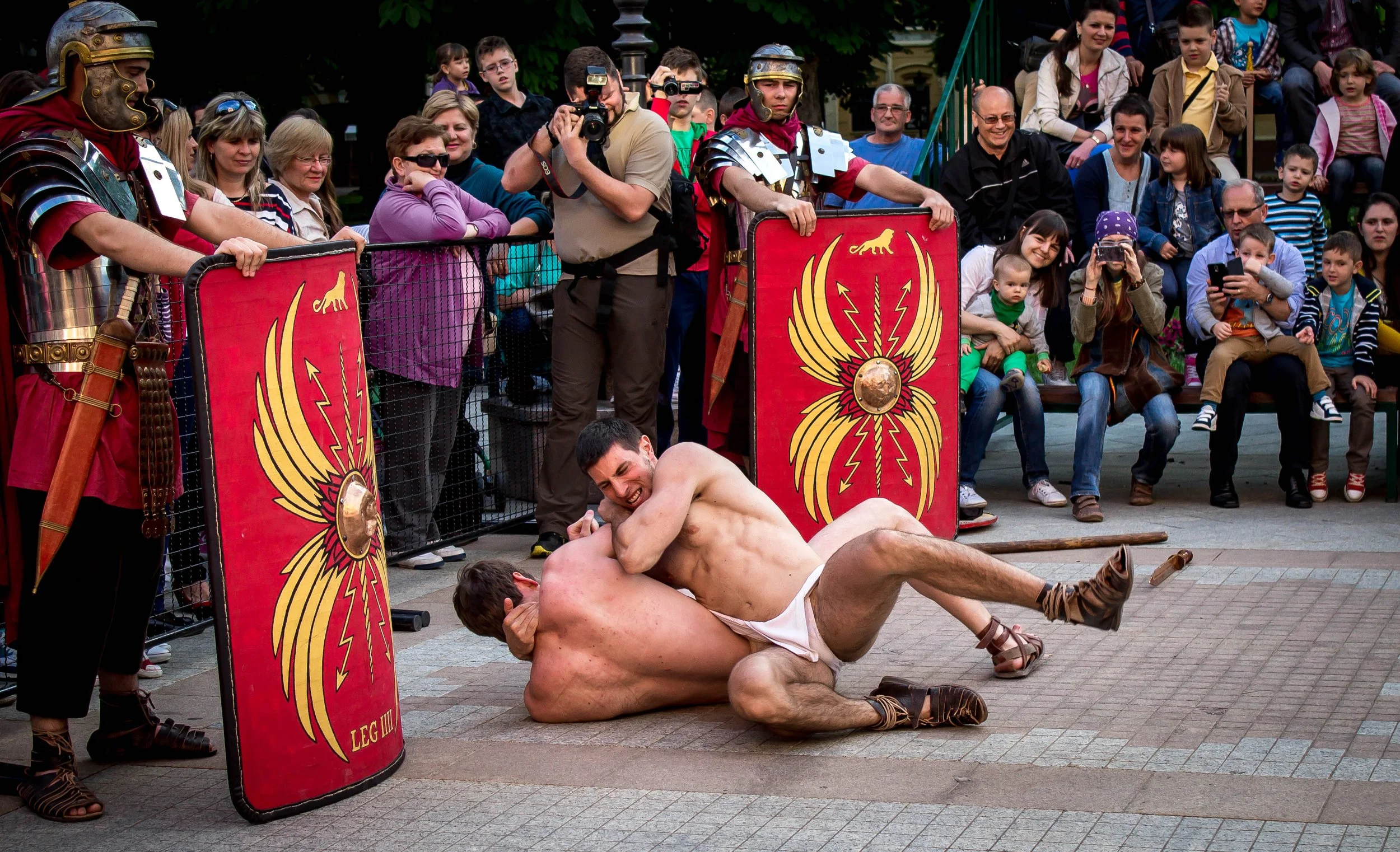 Two men dressed in ancient Roman-style gladiator costumes engaged in a mock fight, one on top of the other on a paved street, surrounded by an audience of adults and children, some taking photos, with Roman-style shields and armor.