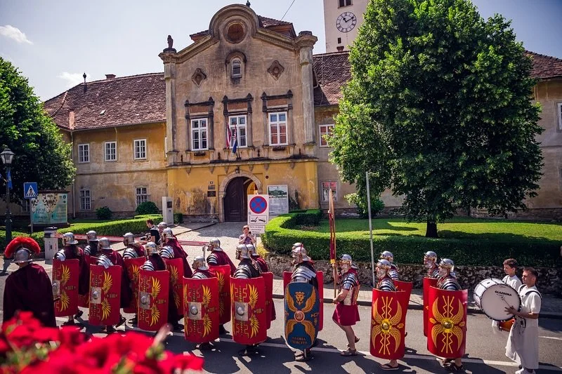 A street parade featuring soldiers in ancient armor, carrying shields with Roman symbols, walking past a historic building with a yellow facade and a green tree in front.
