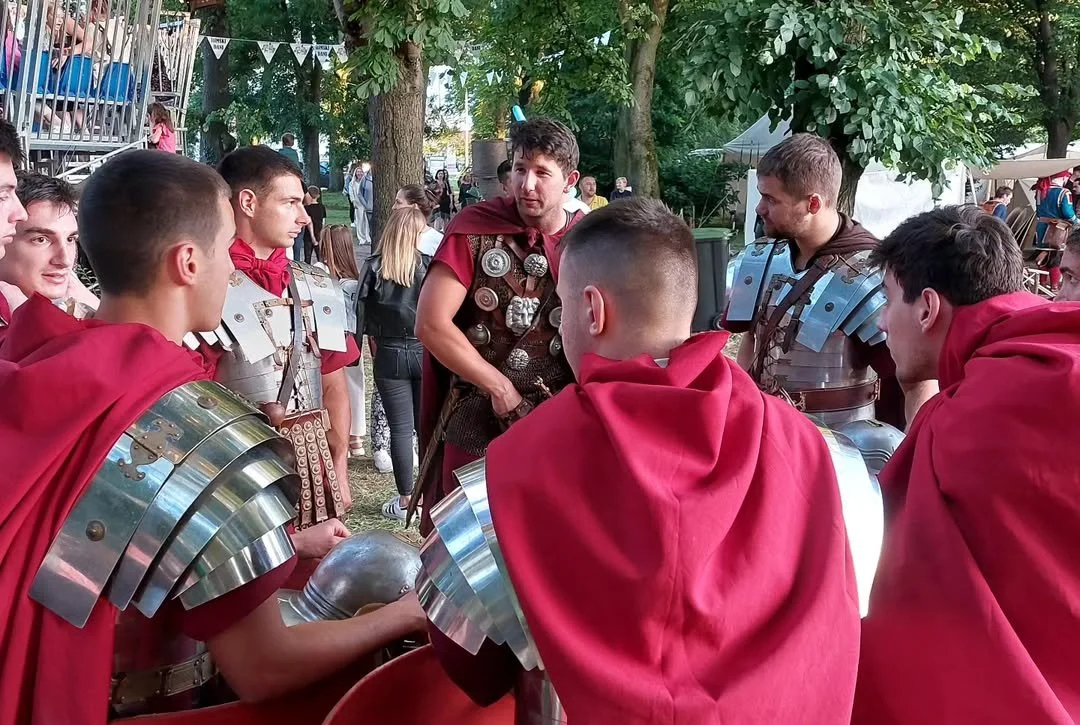 A group of young men dressed as Roman soldiers with armor and red cloaks, gathered around and listening to a man also in Roman costume, in an outdoor setting with trees and spectators in the background.