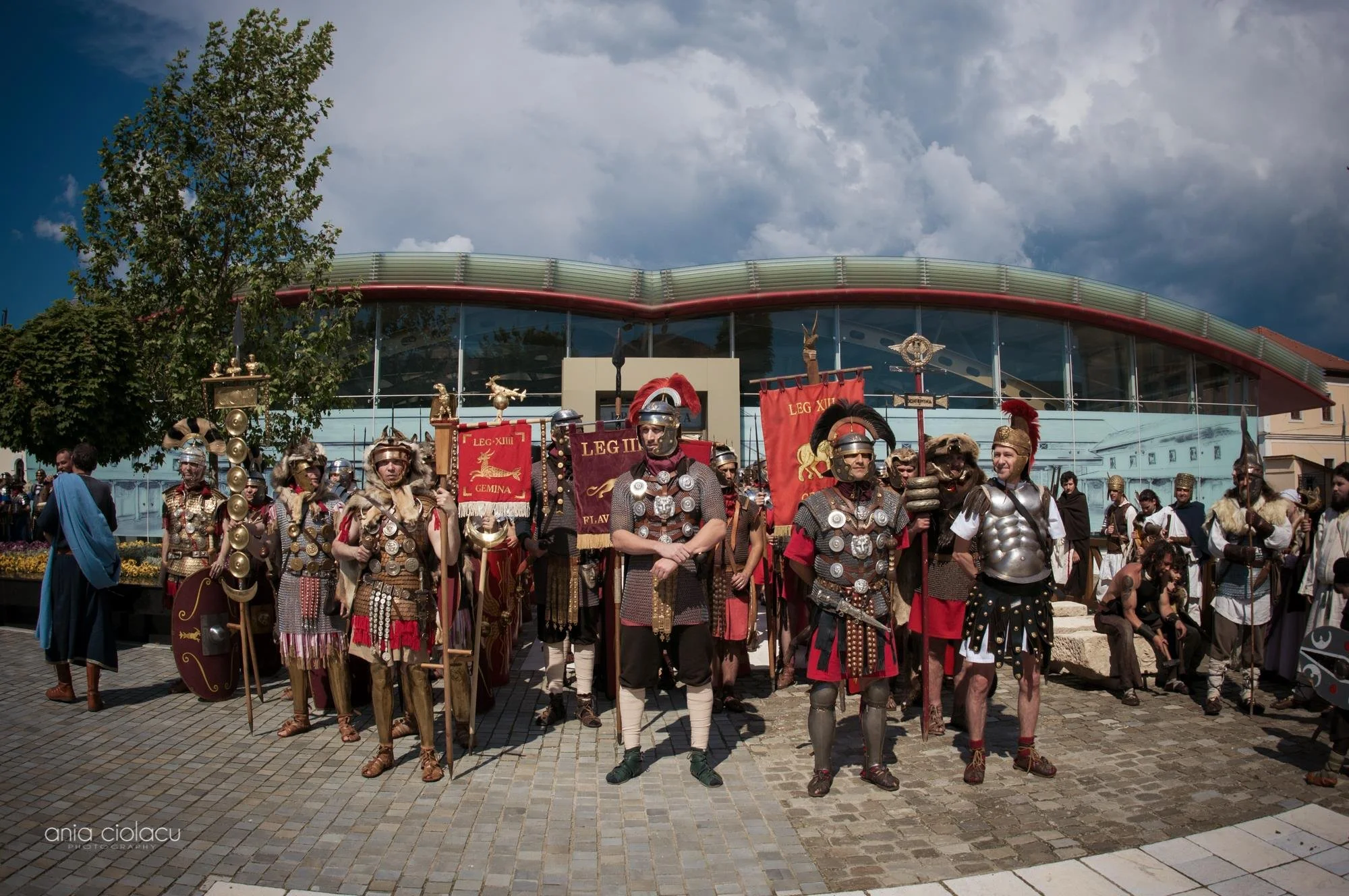 Group of people dressed as Roman soldiers in historical costumes standing in front of a modern building with a curved glass roof, under a partly cloudy sky.