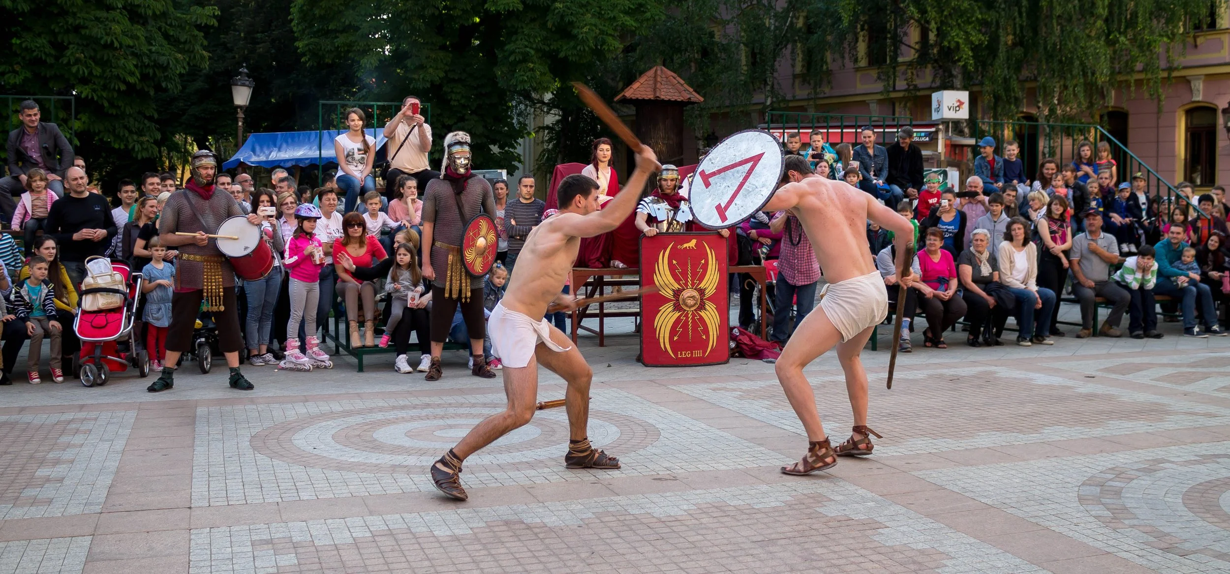 Two men dressed as ancient Roman gladiators fighting with swords and shields in front of a large crowd seated on benches, with some spectators taking photos.