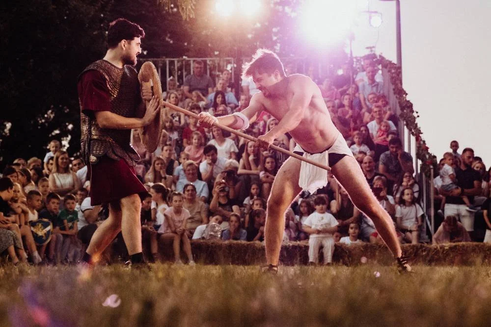 Two men engaged in a reenactment of a gladiatorial combat in front of a large audience, one with a shield and the other with a spear, in an outdoor setting during sunset.